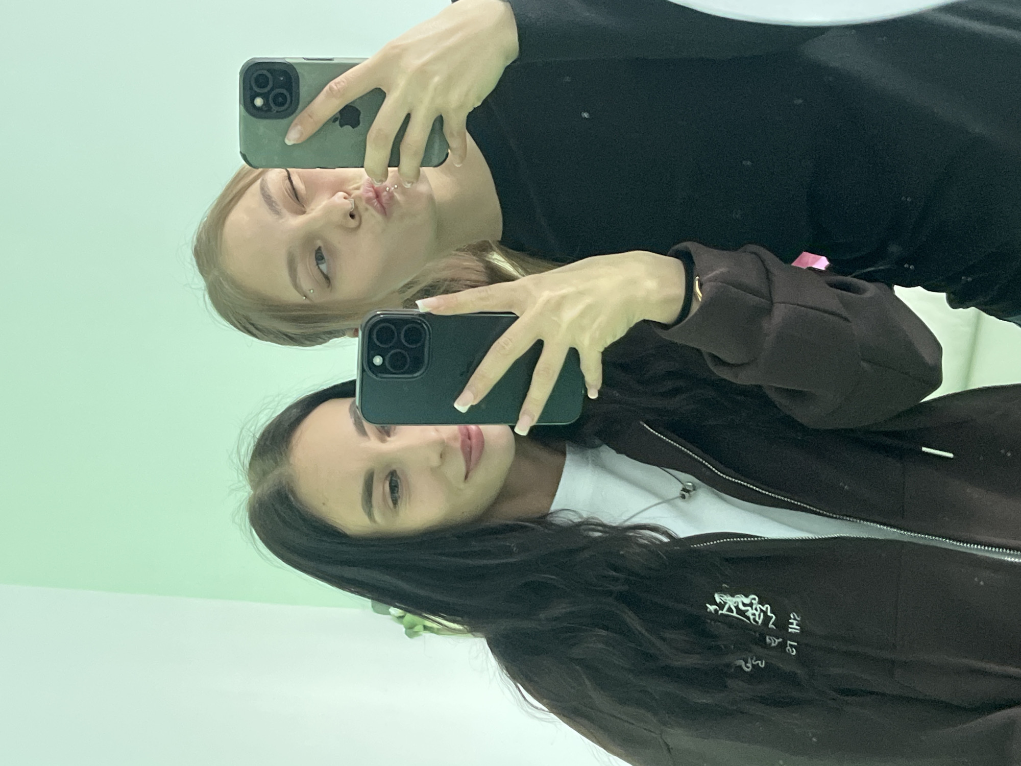 This image, rotated 90 degrees clockwise, captures two young women taking a mirror selfie indoors in Фоки, Russia.
On the left side of the frame (when upright), a woman with blonde/light brown hair is seen from the chest up, wearing a black long-sleeved top. She poses playfully, making a "duck face" or blowing a kiss, and has multiple facial piercings including a nose stud, an eyebrow piercing, and a lip piercing. She holds an iPhone with a dark green case, taking the photo.
To her right and slightly below, the second woman, with long dark brown hair, is also visible from the chest up. She wears a dark brown zippered hoodie over a white t-shirt and looks towards the camera with a subtle, relaxed smile. Her hand, showing manicured nails with French tips, holds another iPhone with a black case.
The background of the mirror reflection is a light mint green color. On the dark brown hoodie, there is a partially obscured, white stylized graphic or logo, which is difficult to read due to mirroring and its angle. The overall scene portrays a casual, friendly interaction between the two women as they capture a moment together.