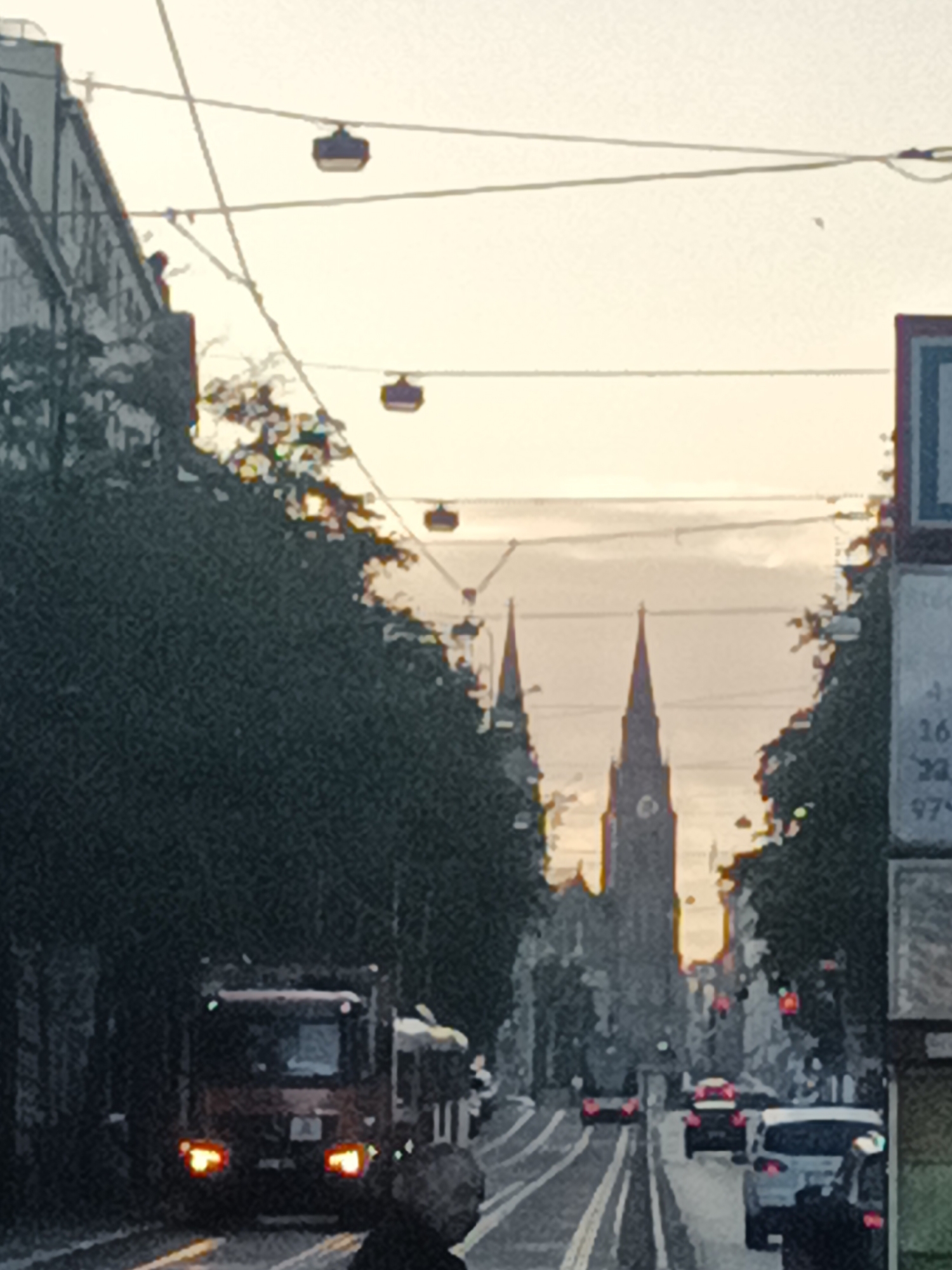 A street scene in Prague, Czechia, features tram lines overhead and a prominent church with two spires in the distance, silhouetted against a pale sky. A tram or similar vehicle is visible in the foreground, with its headlights on. Cars are moving along the street, and trees line both sides, partially obscuring the buildings. The overall impression is of a city street during daylight, possibly in the late afternoon or early evening given the lighting.