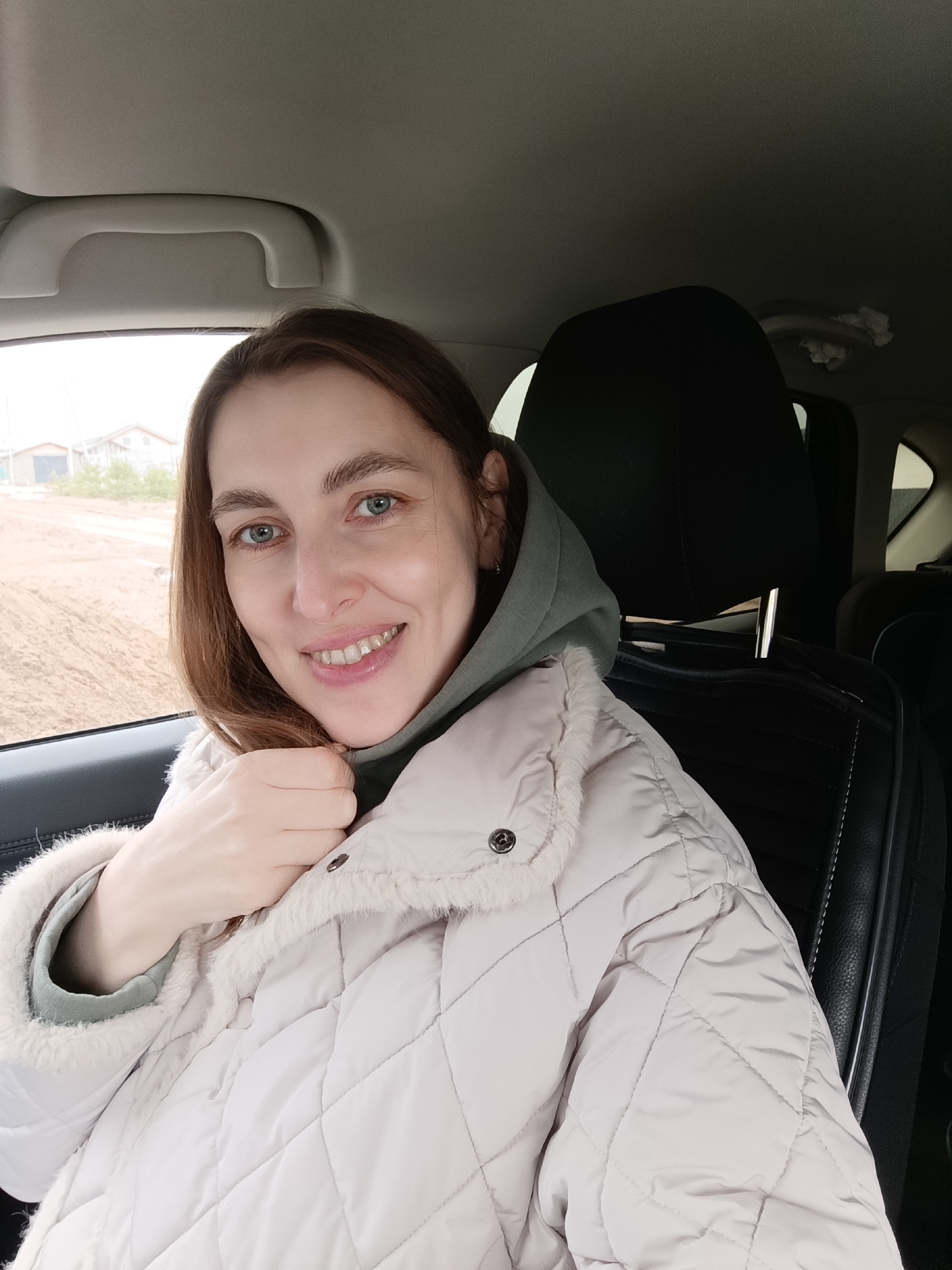 A medium shot depicts a woman sitting in the driver's seat of a car. She is smiling and looking directly at the camera. She is wearing a light-colored, quilted jacket with a furry collar over a hooded sweatshirt. Her hair is light brown, and her eyes are blue.
The car's interior is visible, including the headrest, the door panel, and the ceiling. Through the car window, a blurred landscape can be seen, featuring a dirt road, houses, and possibly other buildings. Based on the information provided, the location is identified as Chaikovsky, Russia.