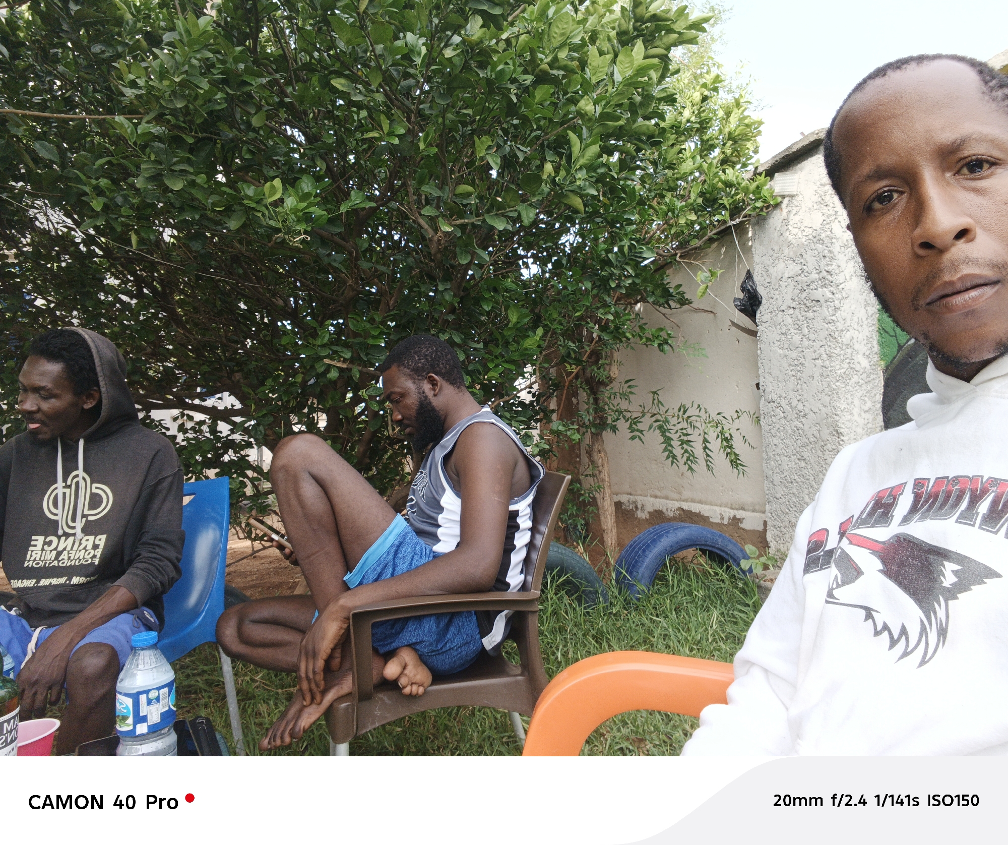 The image captures three African men seated in outdoor chairs. One man wears a grey hooded sweatshirt with a graphic design and blue shorts. Another man, seated in a brown chair, is shirtless with a tank top and blue shorts. The man in the foreground wears a white hoodie with a graphic design. They are seated in a grassy yard, near trees. There is a water bottle on the ground, as well as a blue tire. The camera and image details are listed at the bottom.
