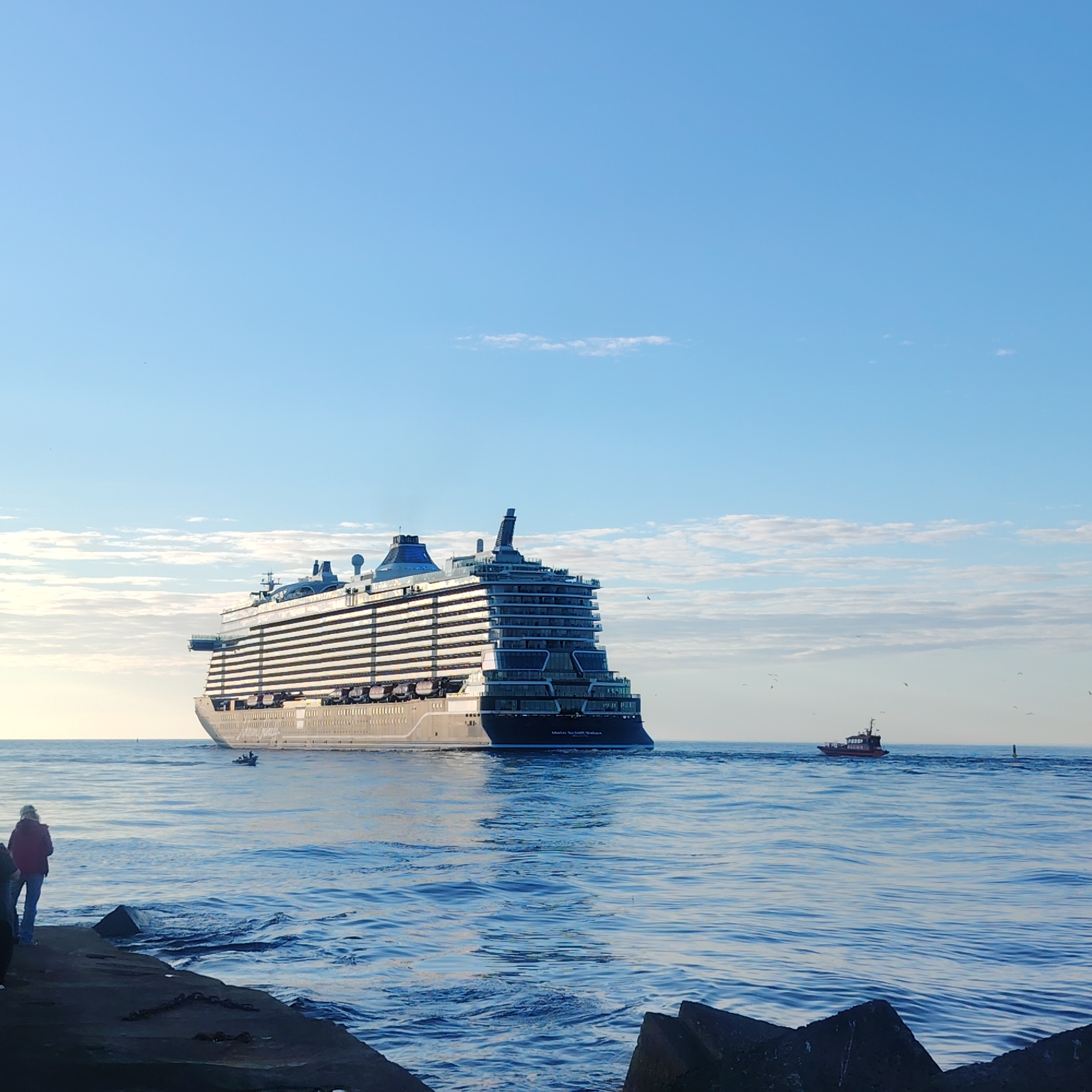 The image shows a large cruise ship on the water, likely entering or exiting the port. The ship is multi-storied and modern, with multiple decks and balconies. A smaller pilot boat is positioned near the cruise ship. The water is blue with some wave action. A person in a red jacket and another person are standing on a concrete breakwater in the foreground. The sky is mostly clear blue with some scattered clouds. The location is likely the port of Riga, Latvia, given the visual cues and the presence of maritime traffic.