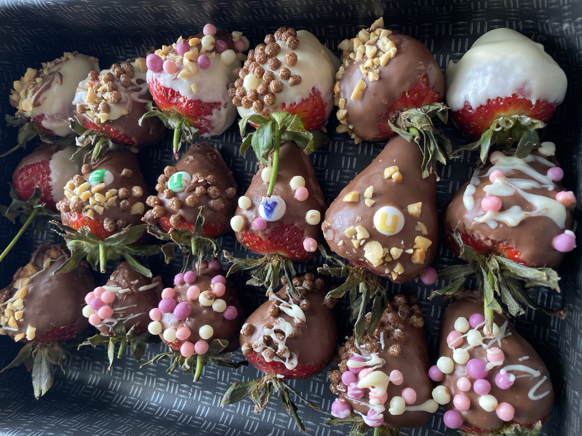 A top-down, close-up shot displays a collection of 18 gourmet chocolate-covered strawberries arranged in a dark, textured box or tray. Each strawberry is dipped in either milk chocolate or white chocolate and adorned with various toppings, creating a visually appealing dessert assortment.
The strawberries feature a mix of dark chocolate and white chocolate coatings. Some are fully covered in milk chocolate, while others are half-dipped in white chocolate, showcasing the vibrant red of the strawberry underneath. The toppings include chopped nuts (likely almonds or peanuts), brown cereal-like crisps or cocoa puffs, and various colorful sprinkles in shades of pink, purple, white, and light green. Many also have delicate drizzles of contrasting white chocolate or milk chocolate on top.
Several strawberries feature small square or circular candy pieces with single letters inscribed on them. Visible letters include a yellow square with a white "U", a green square with a white "E", a light blue-purple square with a white "Y", and another light blue-purple square with what appears to be a white "J" or "T". The stems of the strawberries are still attached, providing a natural green contrast to the rich colors of the chocolate and fruit. The dark, patterned background of the box enhances the presentation of the colorful treats, suggesting they are prepared for a special occasion, gift, or indulgence.