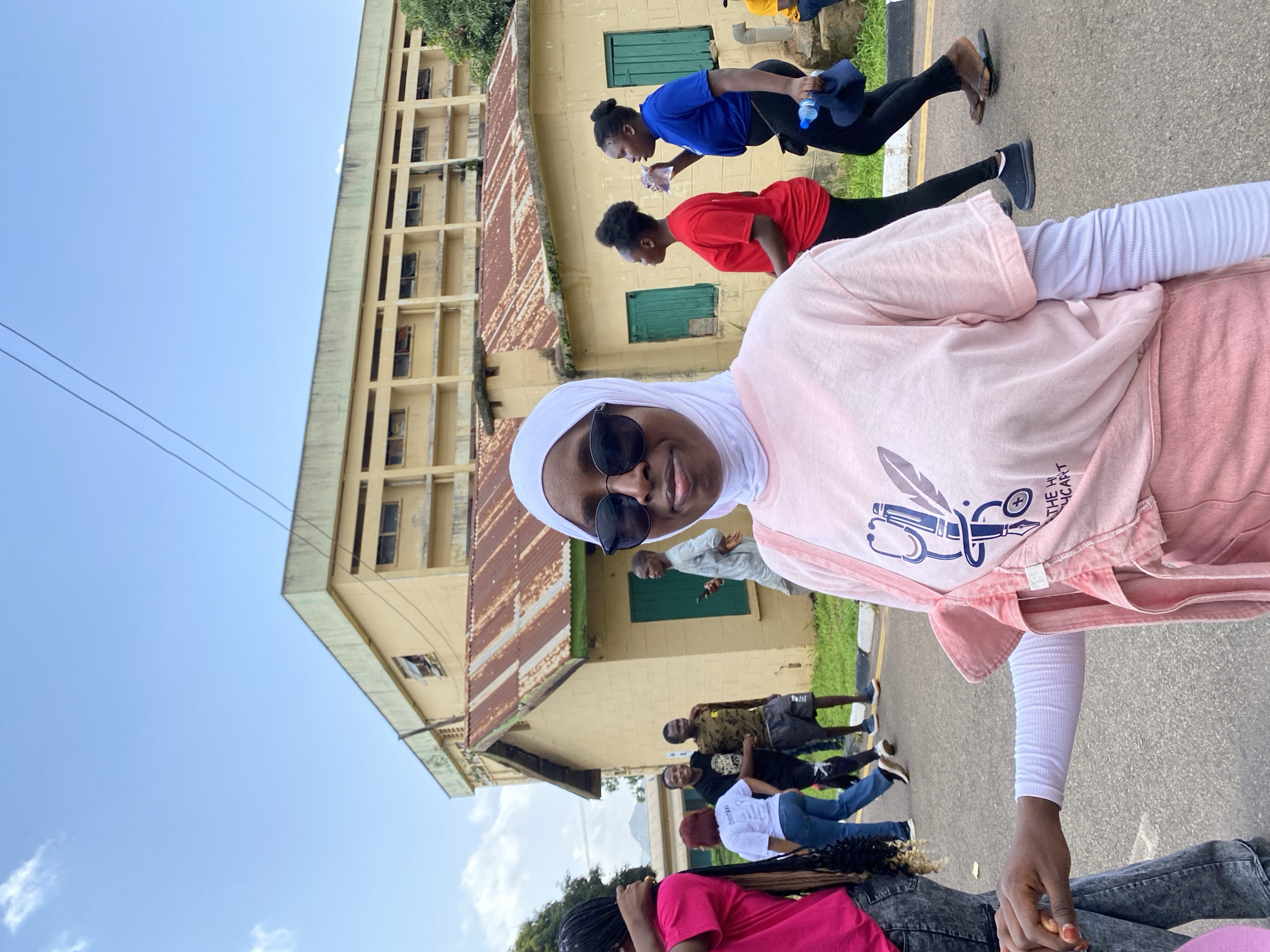 The image depicts a group of young people gathered outdoors in what appears to be a school or institutional setting. In the foreground, a person wearing a pink long-sleeved shirt, a white hijab, and sunglasses is looking at the camera, holding hands with another person. Behind them, several other individuals are visible, some standing and some in motion.
The background features a two-story building with a pale yellow facade and a rusted, corrugated iron roof, suggesting an older structure. There are green-paneled windows on the building. The ground is asphalt, indicating an outdoor paved area, possibly a walkway or courtyard. The sky is visible, with a few scattered clouds, suggesting it is daytime. The overall scene suggests a casual gathering or an organized activity taking place outdoors.
The people are dressed in casual attire. One person wears a blue t-shirt and black pants, another a red t-shirt, and others are in t-shirts and jeans. Some individuals appear to be walking or engaged in an activity. The presence of a water bottle held by one of the individuals might suggest physical activity or a break.
There is text visible on the pink shirt worn by the person in the foreground, featuring a graphic of a quill pen, a stethoscope, and what appears to be a caduceus symbol. Some of the text reads "THE HEALTH CART."
The setting, with the architecture and the light, points to a warm, possibly sunny day. The emotions conveyed are generally neutral to positive, with the person in the foreground offering a slight smile. The location context of Jos, Nigeria, aligns with the architectural style and the appearance of the individuals.