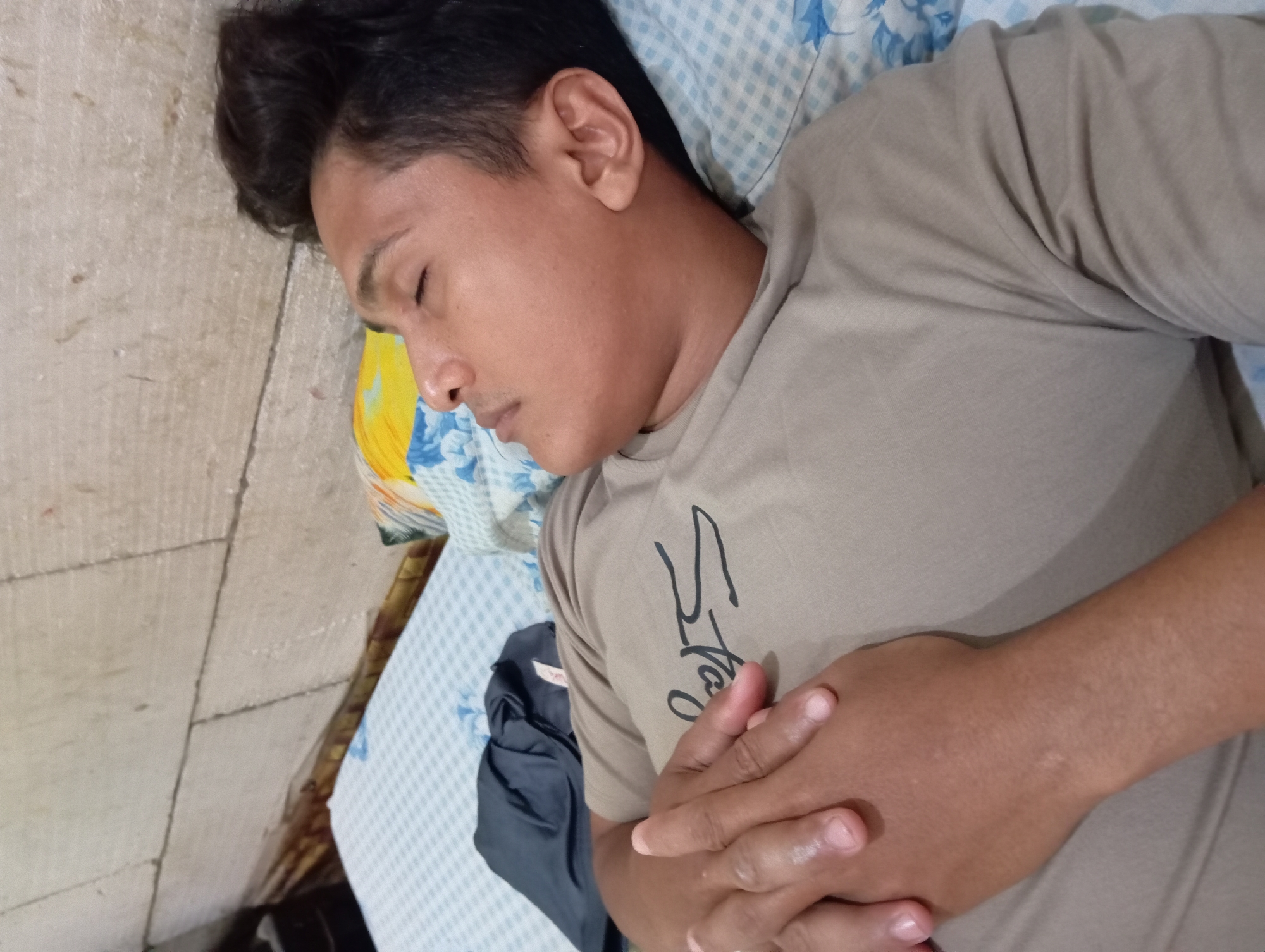 A person with dark hair is lying on a bed with a patterned pillow. The person is wearing a light brown t-shirt and has their hands clasped on their chest. A dark-colored garment is near the person. The bed is positioned on a floor with tile flooring. The location is Baron, Indonesia.