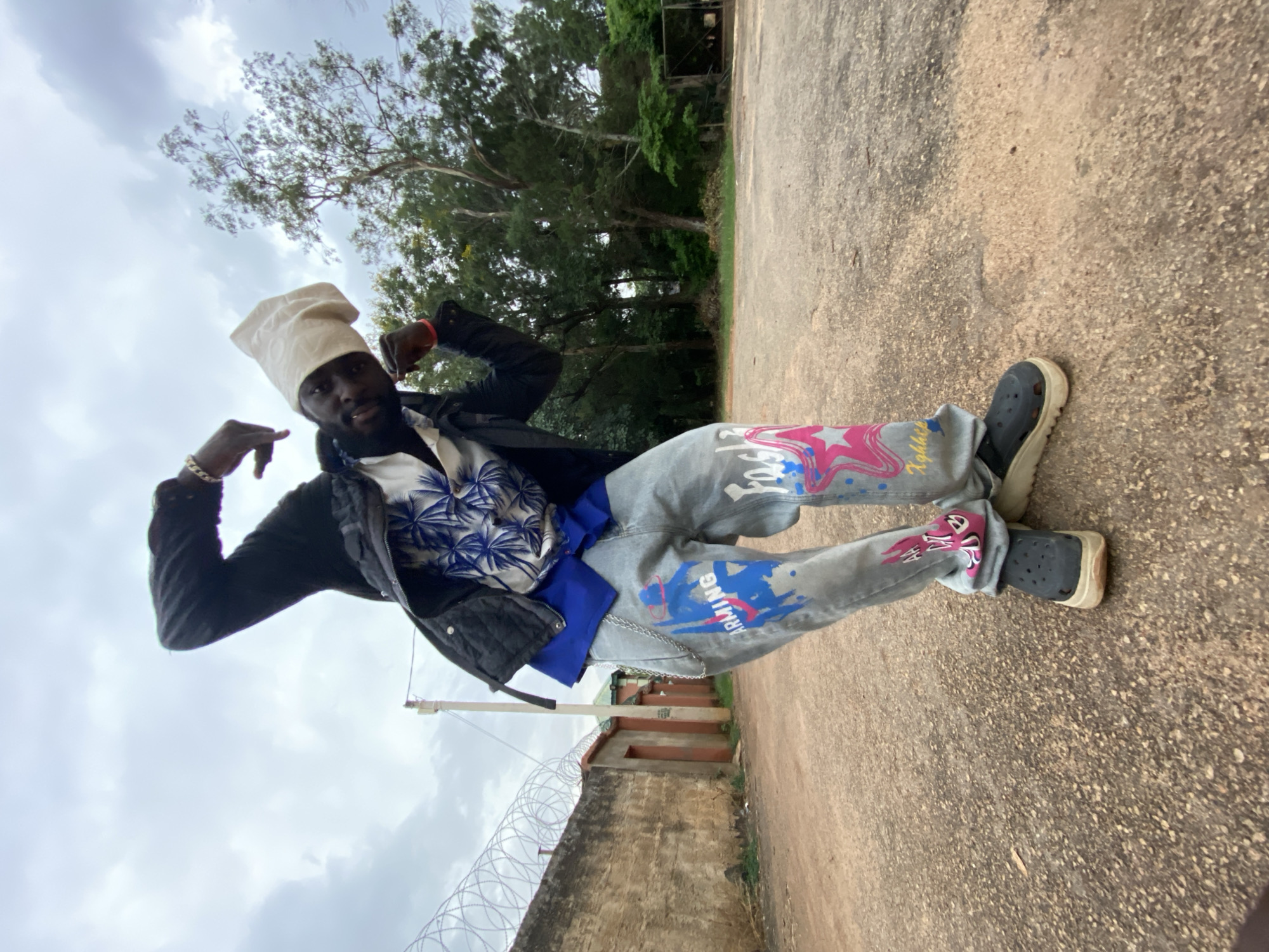 A man is posing outdoors in what appears to be the city of Bukuru, Nigeria. He is wearing a white chef's hat, a blue and white patterned shirt, a black jacket, light gray jeans with colorful graffiti-style designs, and dark-colored Crocs. He is striking a pose with his arms raised. The background includes a concrete surface, trees, a fence topped with barbed wire, and a building. The sky is overcast.