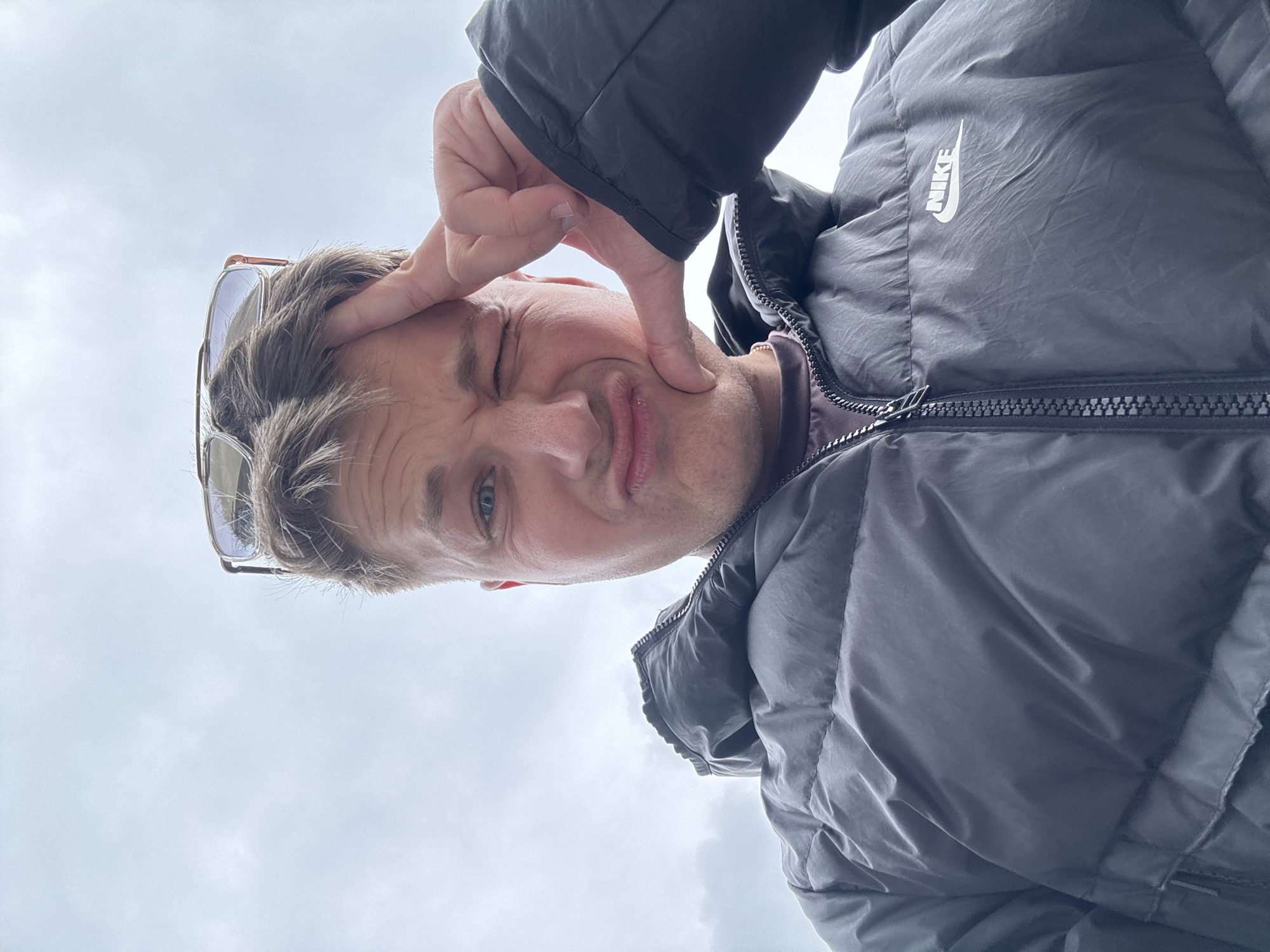A man is pictured from a low angle. He is wearing a dark, quilted Nike jacket. He is making a playful face, winking and pursing his lips while touching his forehead with his fingers. He has sunglasses perched on his head. The background is a pale, overcast sky. Location: Burgas, Bulgaria cannot be determined from the image.