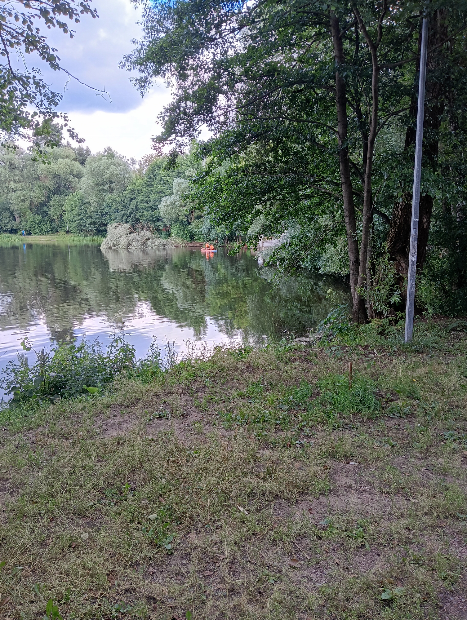 An outdoor scene depicts a natural environment with a body of water and vegetation. The foreground consists of dry, patchy grass and dirt. Beyond the grass, a body of water, likely a lake or pond, is visible, reflecting the surrounding trees and sky. Two small orange buoys are floating on the water, suggesting recreational activity or hazard marking. A fallen tree trunk, partially submerged, lies near the buoys. Dense green foliage and trees line the far bank of the water. In the background, a single, thin grey pole stands upright on the right side of the frame, possibly a lamp post or utility pole. The sky is overcast with patches of blue. The overall impression is of a park or natural area. The image was taken in Kratovo, Russia.