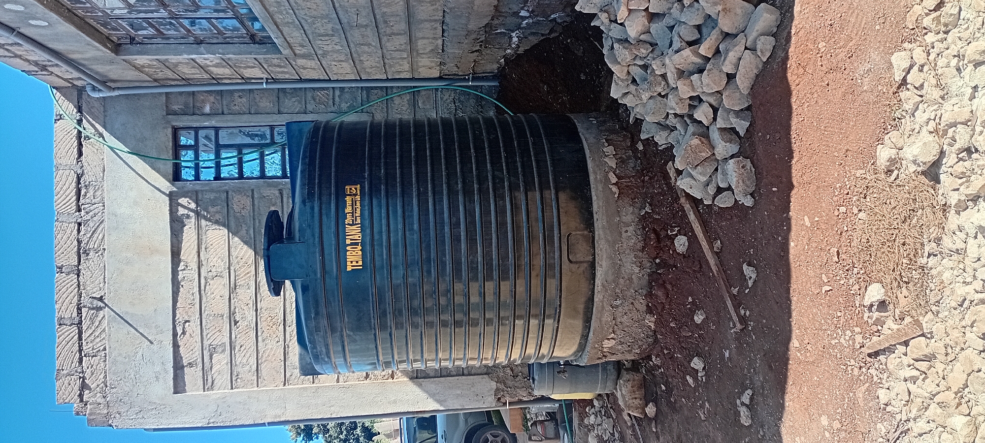 A black, ribbed, cylindrical water tank with the label "TEMBO.TANK 20yrs Warranty Save Water, Save Life." is situated on a circular concrete base in front of a building under construction. A green hose is connected to the top of the tank and runs up the side of the building. The building's exterior walls are constructed with light-colored, textured bricks. A blue-tinted window is visible on the building. To the left of the tank is a grey plastic barrel. The ground is covered with red soil, scattered rocks, and some building materials. A small portion of a car is visible on the far left of the image. The sky above is clear and blue.