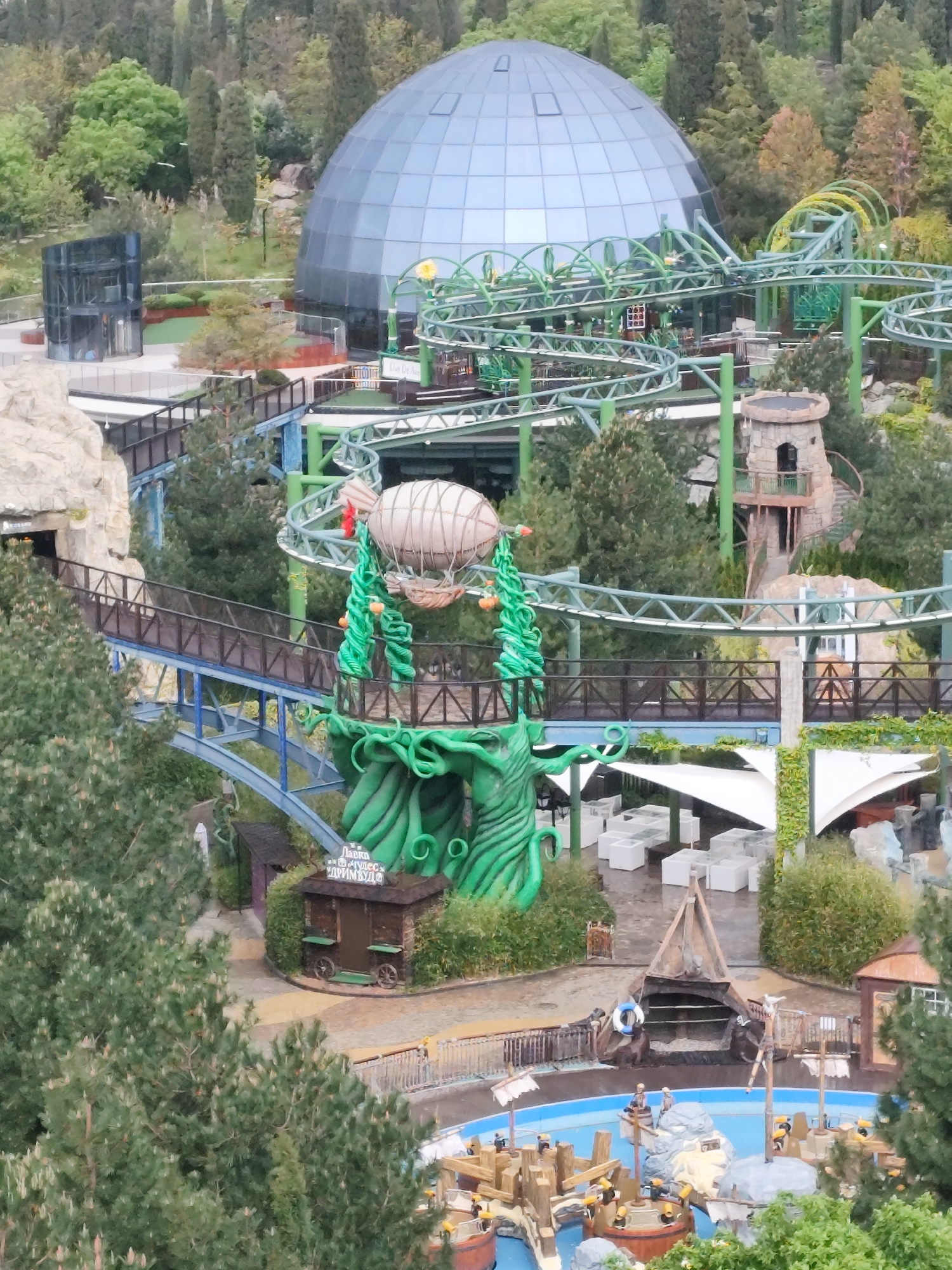 The image captures a high-angle view of what appears to be an amusement park or themed entertainment area. Prominently featured is a large, silver dome-shaped structure, possibly a planetarium or covered attraction. A green roller coaster track weaves through the scene, passing by a decorative air balloon mounted on a green, root-like structure. A small castle-like tower with an outdoor staircase is visible amongst the trees. Below the roller coaster, there is an outdoor dining area with white tables and chairs under white canopies. A water feature with boats or rafts populated with figures are situated in the lower portion of the image, alongside various themed structures. The environment is heavily landscaped with various trees. The location is Симеизский поселковый совет, Ukraine.
