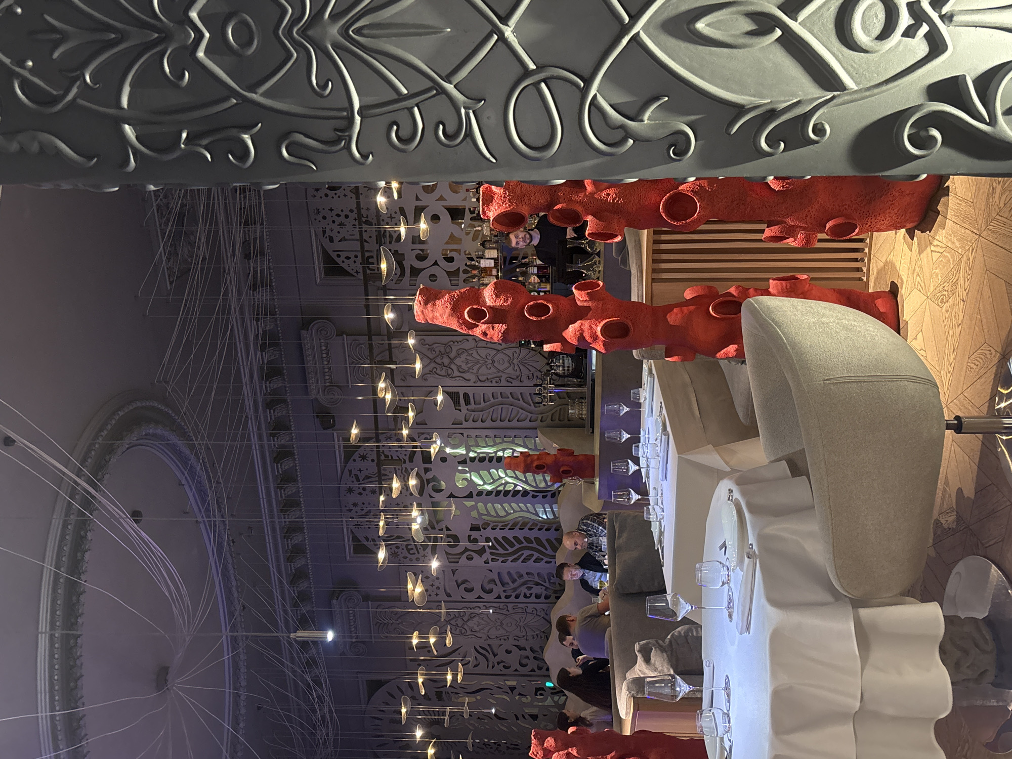 The image depicts the interior of a high-end restaurant or bar located in Krasnoyarsk, Russia. The scene is illuminated by artificial lighting, suggesting an evening setting.
The foreground on the right features a portion of a large, grey, decorative column adorned with intricate, raised swirling patterns. To the left of this column, three prominent, tall, irregularly shaped sculptures, colored a vivid red, stand on the wooden parquet floor. These sculptures have multiple circular perforations of varying sizes. The floor exhibits a parquet pattern of light and dark wood tiles.
In the midground, multiple round dining tables are visible, draped with crisp white tablecloths. Each table is meticulously set with white plates, folded white napkins, and an assortment of wine glasses and water tumblers. A light grey upholstered swivel chair is positioned beside one of these tables in the immediate foreground.
Further into the midground, a group of approximately six to eight individuals are seated at a booth and table arrangement. These diners are engaged in conversation, with one male individual looking down at a mobile device.
The background reveals an elaborately decorated space. The walls feature extensive ornamental panels with repeating patterns, some appearing to be backlit, creating a layered, textured effect. The ceiling boasts a large circular molding with numerous thin, white cables extending from a central point, connecting to various small, suspended light fixtures that emit a warm glow. Additional clusters of hanging light fixtures, featuring multiple flat, leaf-like elements, are distributed across the ceiling area.
A bar area is visible in the background, behind one of the red sculptures. A male bartender with a beard, wearing a dark shirt, stands behind the bar, which displays various bottles of spirits. The overall environment suggests an upscale, modern, and artfully designed dining establishment.