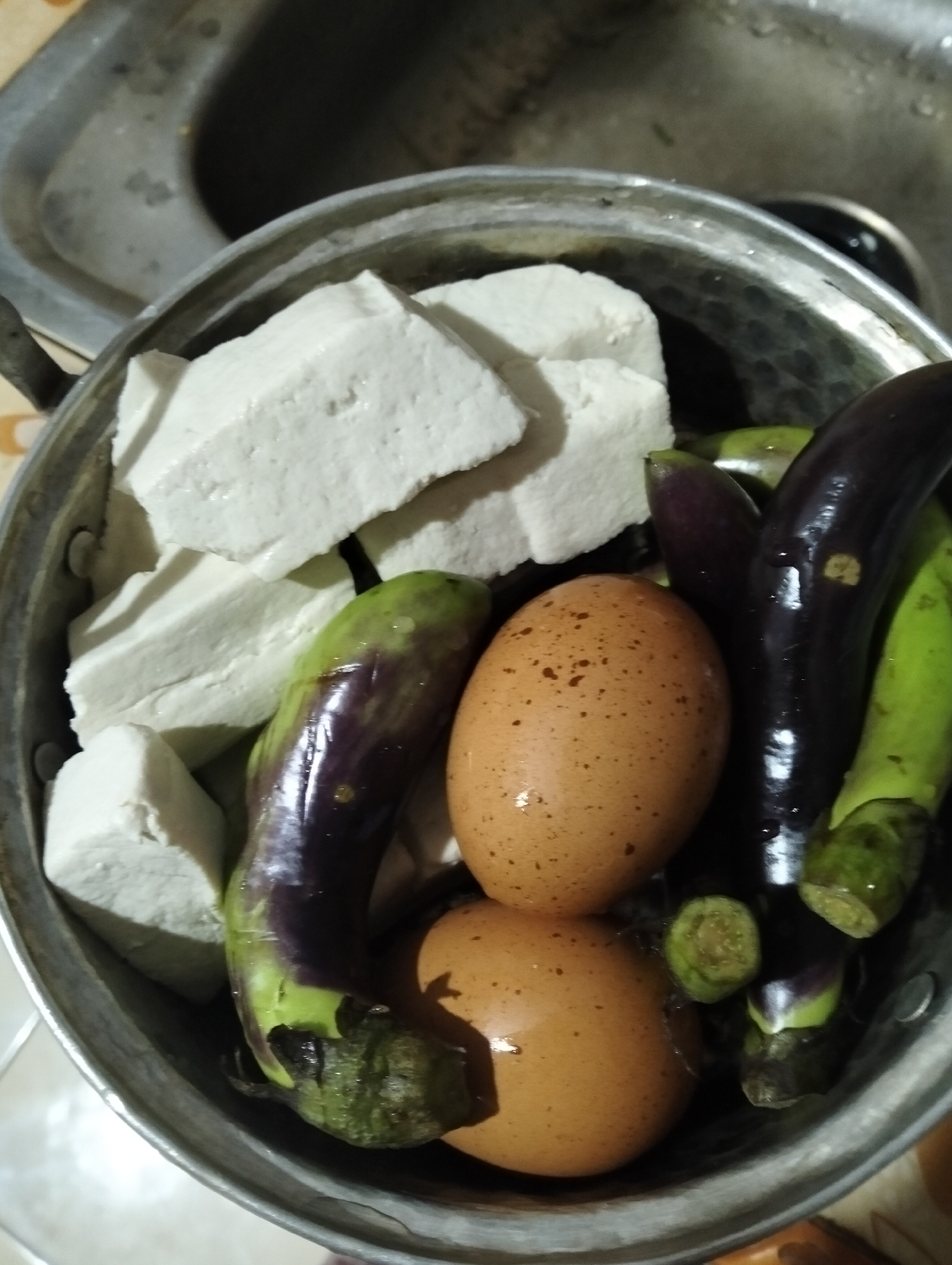 In Muncar, Indonesia, a metal pot is filled with various ingredients. The pot contains several rectangular blocks of white tofu, two brown eggs with speckled shells, and several small, dark purple eggplants with green stems. The pot is placed on a countertop near a stainless steel sink. The lighting suggests an indoor setting, possibly a kitchen.