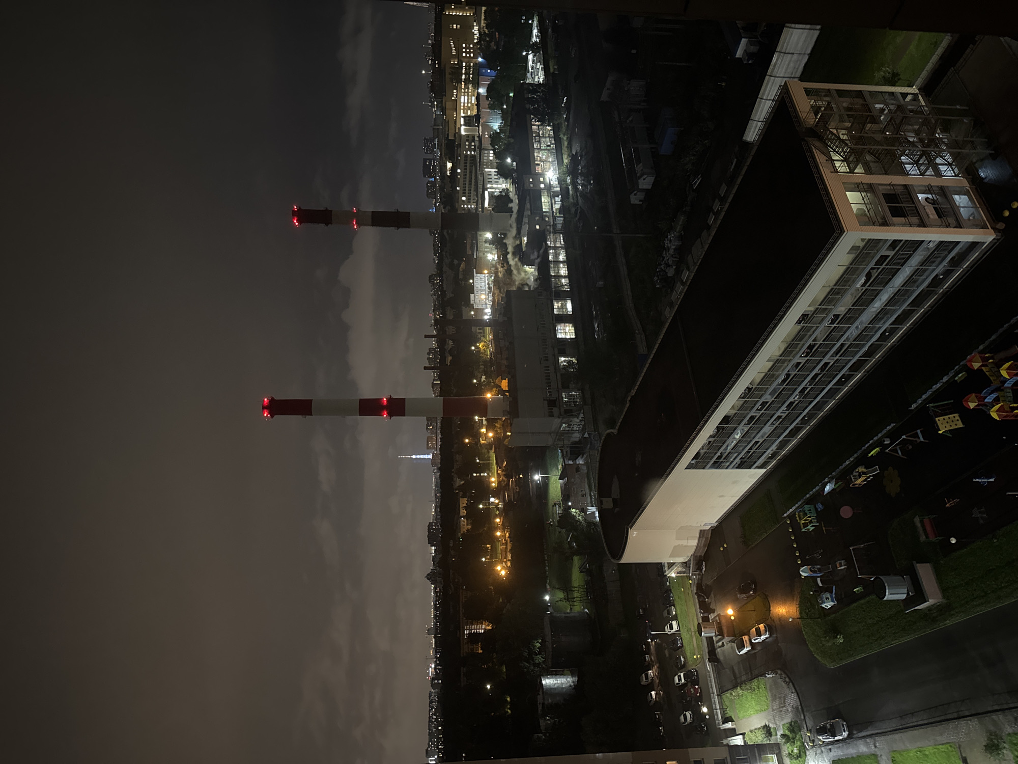 The image is an aerial view of an industrial area and residential complex at night in Sinyavino, Russia. Two tall, red and white striped industrial chimneys dominate the background, their tops illuminated by red lights. A long, multi-story building with a flat roof and external staircases is in the foreground, showing lights on in some of its windows. Adjacent to this building is a dark, wet playground area with various equipment. A parking lot with several cars is visible to the left of the playground, and further in the background, there is a cityscape with numerous illuminated buildings and a tall, slender tower in the distance. The sky is overcast and dark.