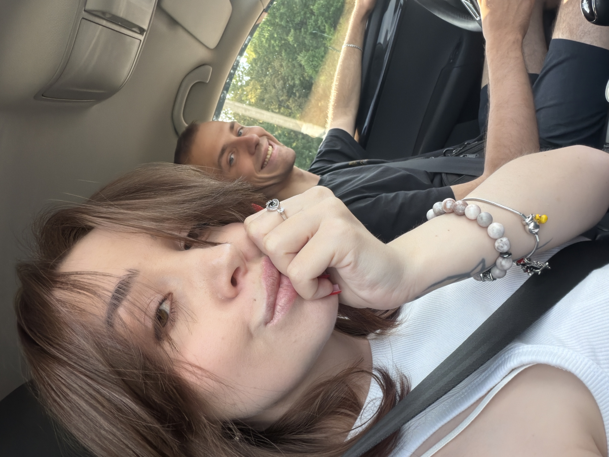 The image shows two people in a car. The person in the foreground is a young woman with brown hair and a white tank top, her hand is touching her mouth. She is wearing a silver ring with a diamond and a bracelet with several round, light-colored stones and charms. The person in the background is a young man with short brown hair, wearing a black t-shirt. He is smiling and looking towards the camera. He appears to be driving. The scene is shot from inside the car, with the outside visible through the windows. Sunlight is streaming in, suggesting it is daytime. The background shows trees and foliage, indicating they are likely driving through a rural or suburban area. The overall mood appears casual and happy. There is no visible text in the image.
