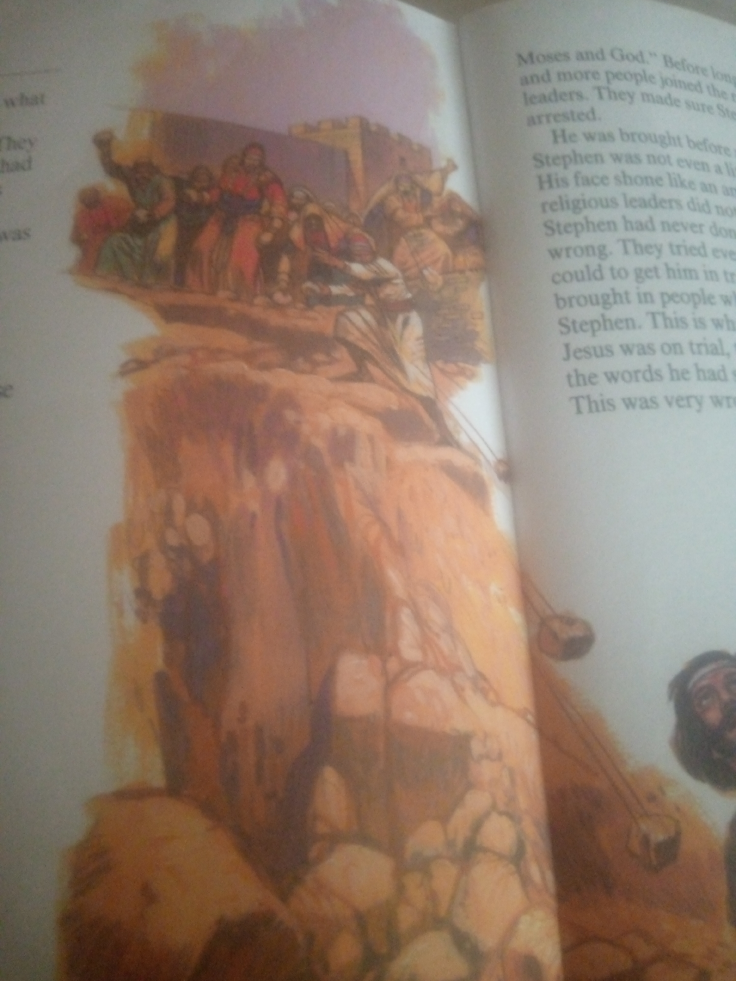 The image is a close-up, slightly blurred photograph of an open book. The left page displays an illustration depicting a group of people on a rocky, elevated surface. Some figures are gathered together, and one appears to be falling or being pushed. There is a suggestion of architecture in the background. The right page contains text, appearing to be from a religious or historical narrative, with the heading "Moses and God." The text discusses a person named Stephen, his trial, and religious leaders. The overall impression is a page from a book with an accompanying illustration, likely depicting a biblical scene, possibly the stoning of Stephen, given the text. The image does not contain any information or visual cues that suggest a location in Jauro Yinu, Nigeria.