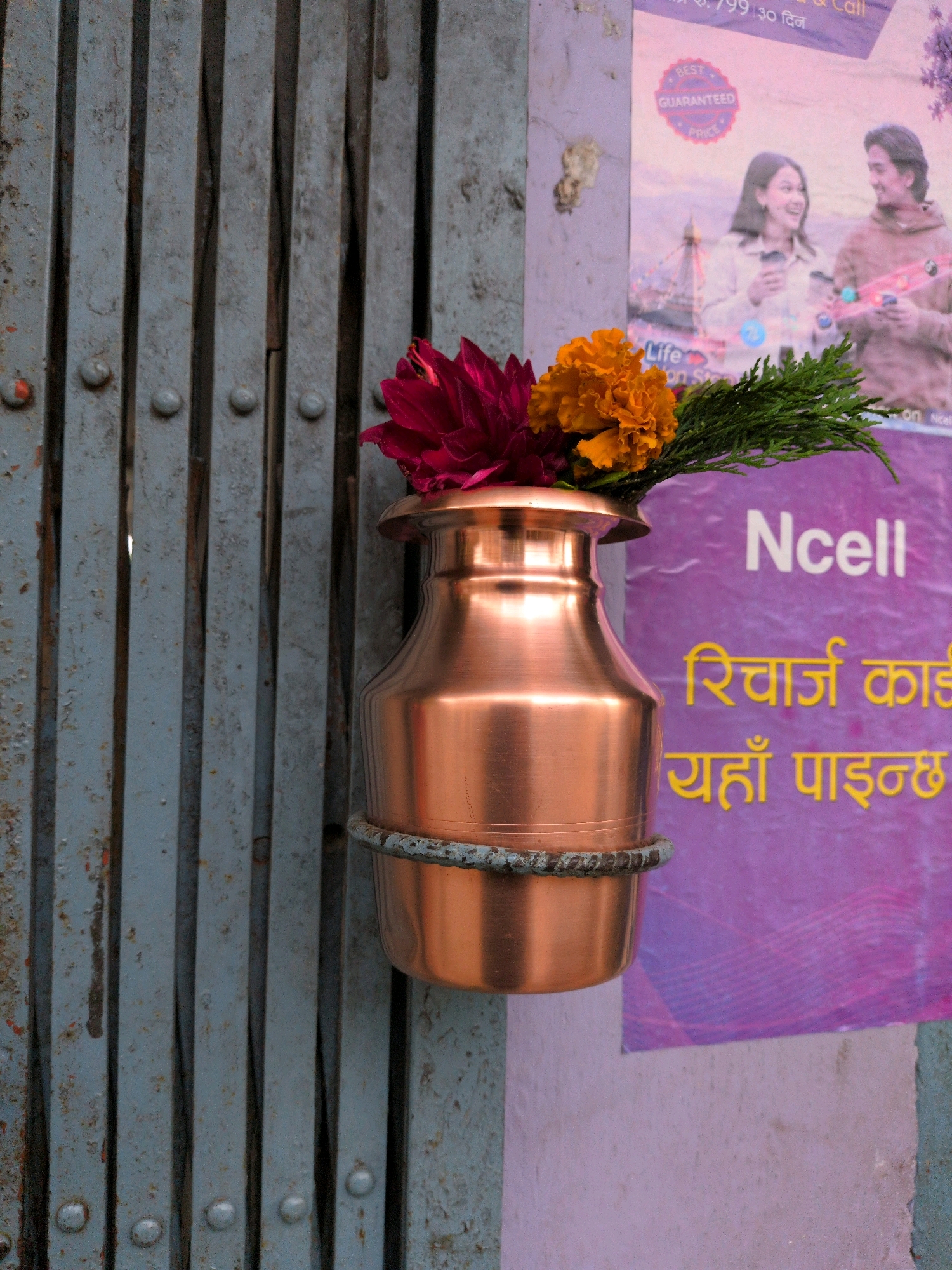 A copper pot filled with flowers is hanging on a weathered grey metal gate. The pot is ornate with a band around its neck, and it contains a vibrant bouquet of red and orange flowers, along with green foliage. Behind the gate, a colorful poster is visible, featuring images of people and text in a language that appears to be Nepali. The poster is predominantly purple and pink, with white and yellow lettering. The background suggests an urban environment, possibly in Kapurkot, Nepal, given the script on the poster. The lighting indicates it is daytime.