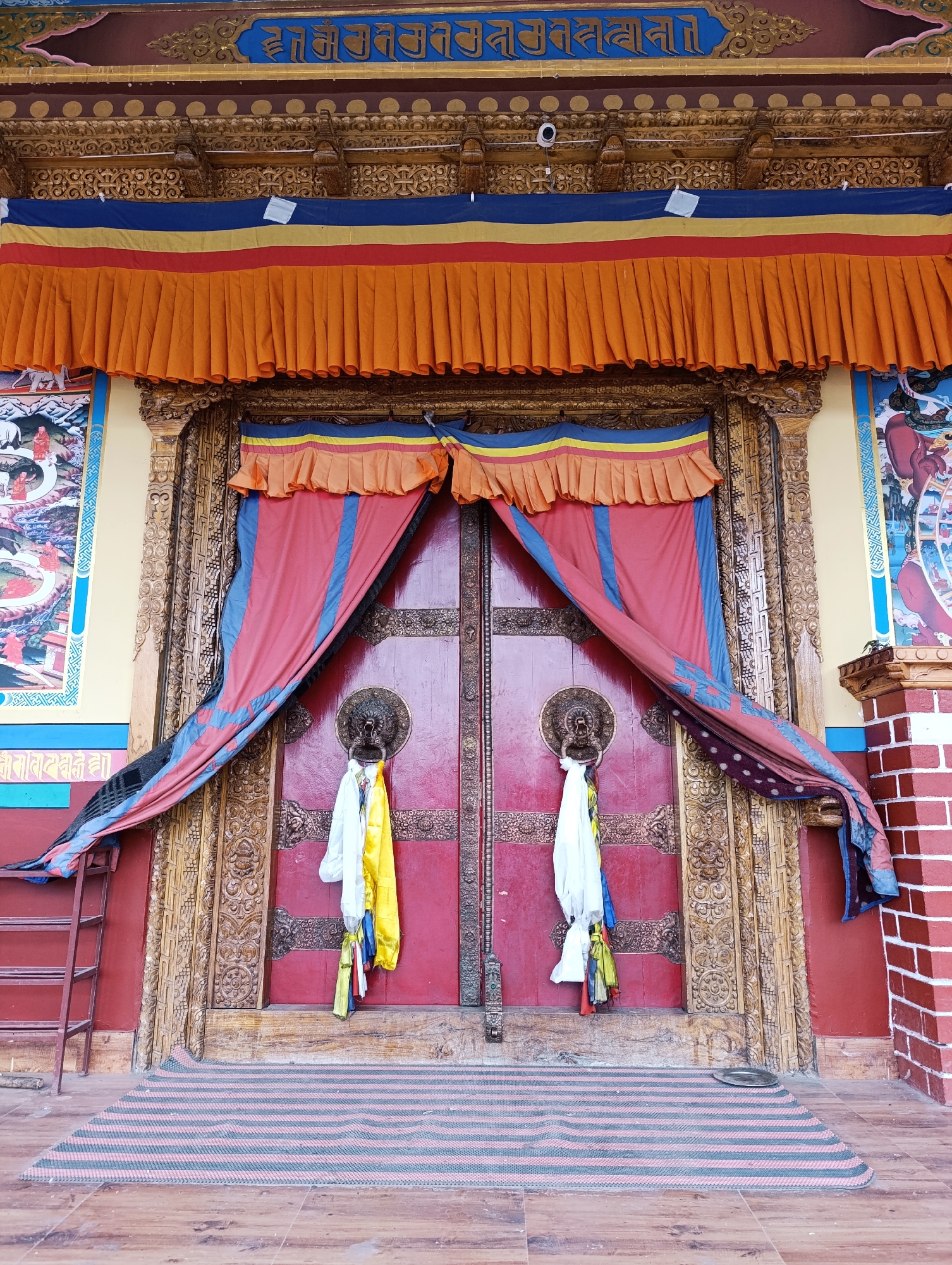 The image depicts the ornate entrance to a building, likely a monastery or temple, in Leh, India. The focal point is a large, red double door adorned with intricate golden carvings along its frame and sides. Two prominent circular brass door knockers, shaped like animal heads, are centered on each door. Draped over the doors are vibrant, pleated fabric curtains in shades of red, blue, and yellow. Hanging from each door knocker are white and yellow prayer flags. Above the door, the building's facade features decorative elements, including gold detailing and blue text in what appears to be a Tibetan script, along with a row of orange, red, yellow, and blue fabric banners. To the left of the entrance, a ladder leans against a wall, and to the right, a wall constructed of red bricks is visible. A striped rug, predominantly red and green, lies on the wooden floor in front of the doors. On the walls flanking the entrance, large colorful murals or paintings are present. The overall scene suggests a place of cultural and religious significance.