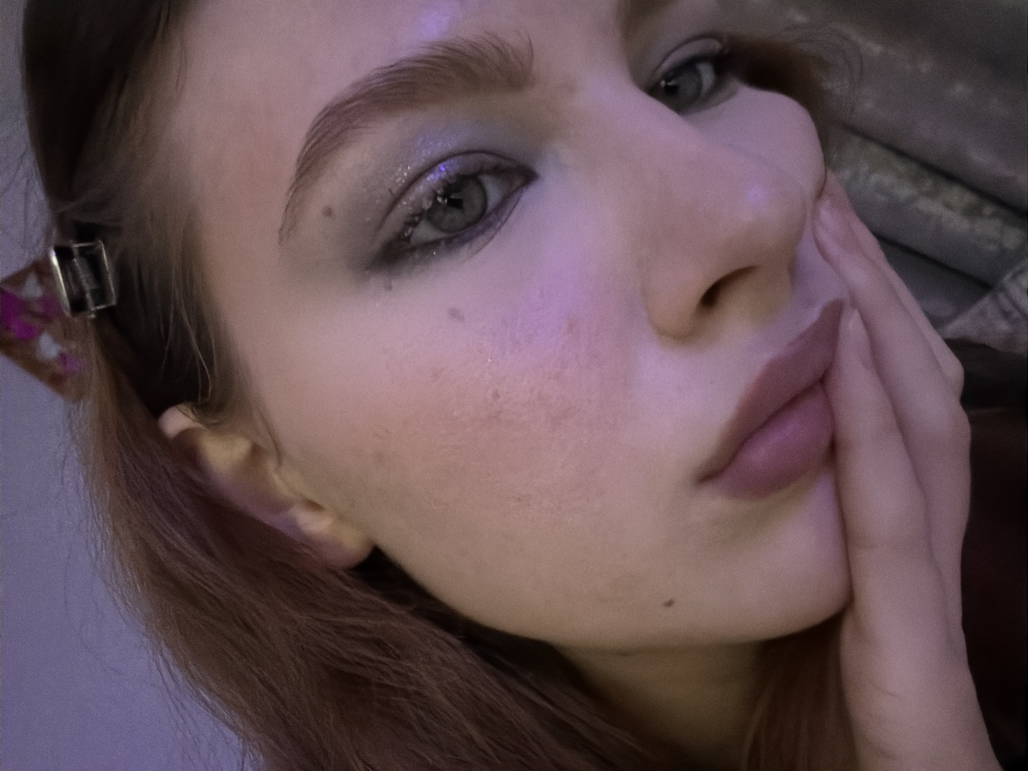 A close-up, low-angle view shows a young woman with fair skin and light brown hair. She is looking slightly downward and to the right. Her left hand is raised to her face, with fingers partially obscuring her cheek. She has light-colored eyes and is wearing eye makeup with some glitter. Her lips are a soft pink color. A hair clip is visible in her hair near her ear. The background is soft and out of focus, with hints of fabric. The scene does not obviously depict anything specific to Noviy, Russia.