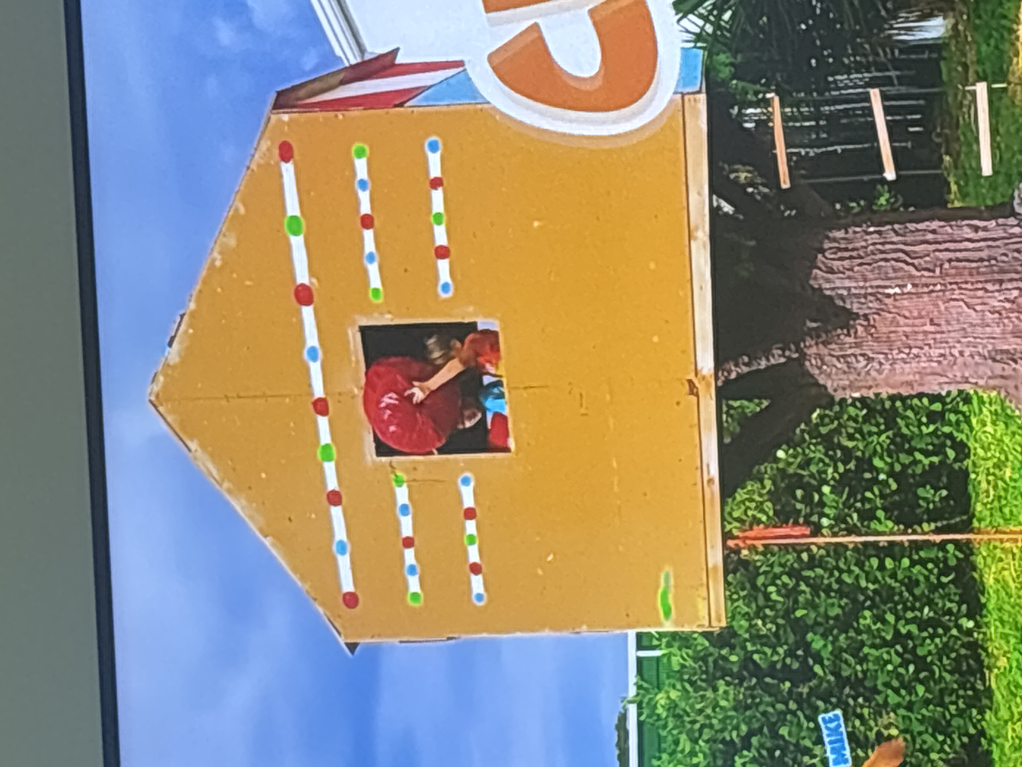 The image shows a child's playhouse resembling a gingerbread house, mounted on a tree. The house is painted yellow and decorated with lines of red, blue, and green dots. A window in the house reveals two children. One child, wearing a blue shirt, is partially visible, while another child, wearing a red shirt, is holding a large red balloon. A ladder made of wooden planks is attached to the side of the tree, leading up to the playhouse. In the background, there is lush green foliage and a bright blue sky. The scene appears to be outdoors, likely in a backyard or park. On the left side of the image, a blue sign with white text reads "MIKE". A large white and orange letter "C" is visible on the right side of the frame. The overall setting suggests a playful and joyful environment. There is no information in the image that places the scene in Dubai, United Arab Emirates.