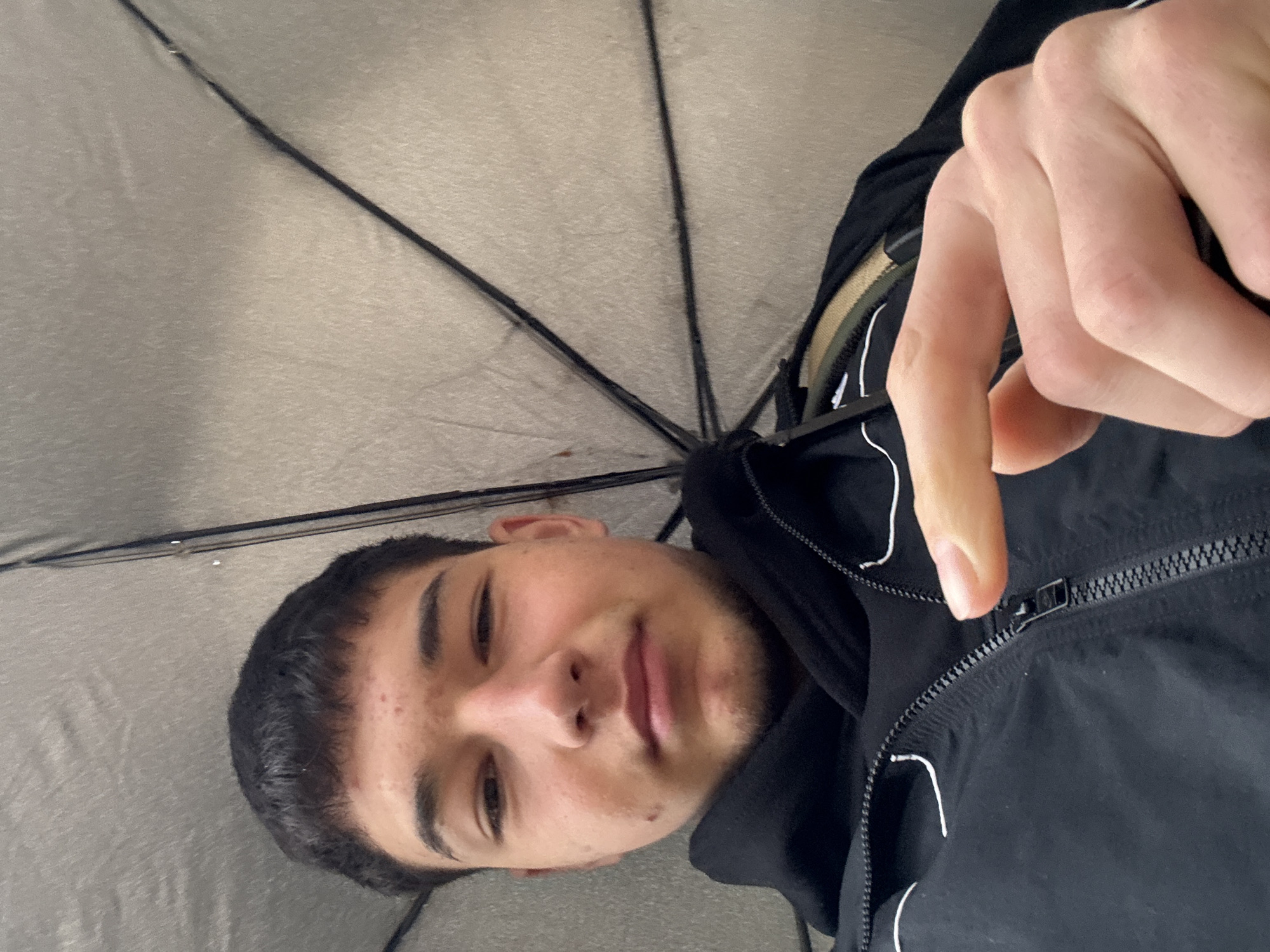 A person is looking upwards, standing under an open umbrella. The person has short, dark hair and appears to be wearing a black jacket with white trim. Their right hand is near the zipper of the jacket. The umbrella is silver or gray and has multiple spokes visible. The specific location in Zlatograd, Bulgaria is not identifiable from the image.