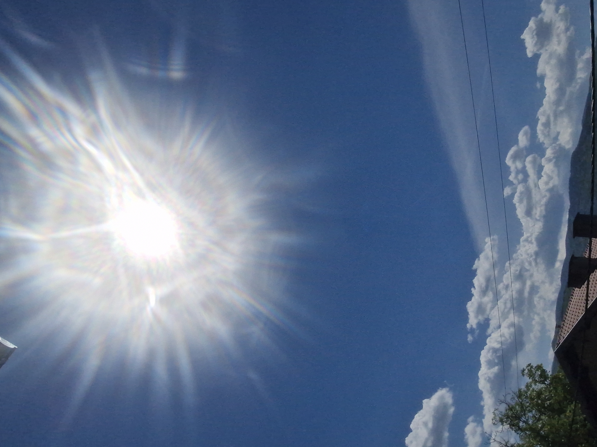 The image is a low-angle shot of a bright, sunny sky in Hrabrino, Bulgaria. The sun is visible in the upper left of the frame, with lens flare emanating outward. To the right, fluffy white clouds are visible, aligned vertically. A few power lines run parallel to the clouds, extending from the top of the frame. The corner of a building with a red tiled roof is visible at the bottom right. Green tree branches and leaves extend from behind the building. The sky is blue.