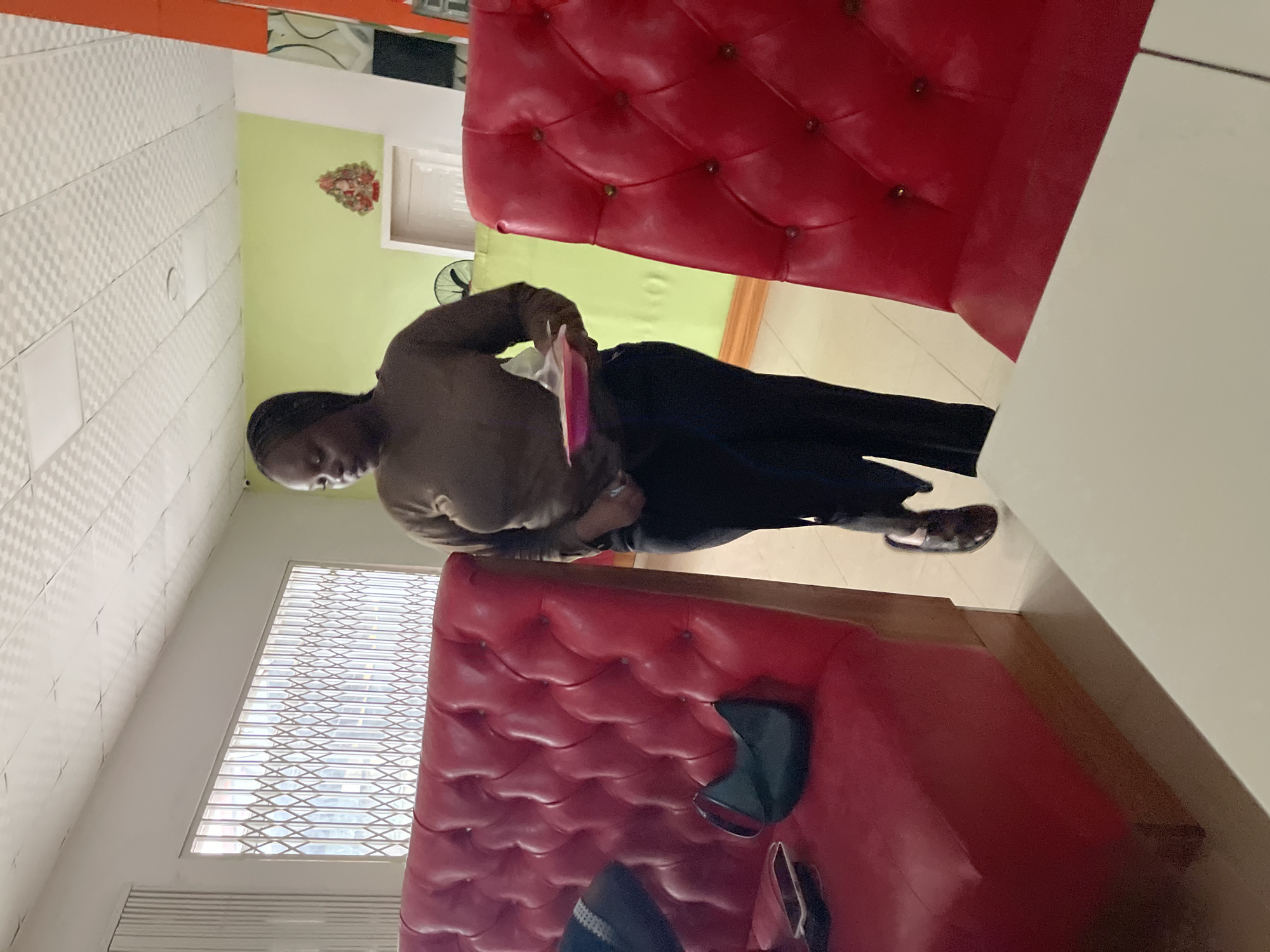 The image shows a woman in a brown long-sleeved shirt and black pants standing in what appears to be a restaurant. She is holding a pink plate. To her left, a red tufted booth is visible with a dark green purse and a phone on it. To her right, another red tufted seat is visible. In the background, a light green wall and a white ceiling with a textured pattern are present. A window with a metal grate is also visible on the left side of the image. The overall scene is indoors and brightly lit. The location is identified as the city of Jos, Nigeria.