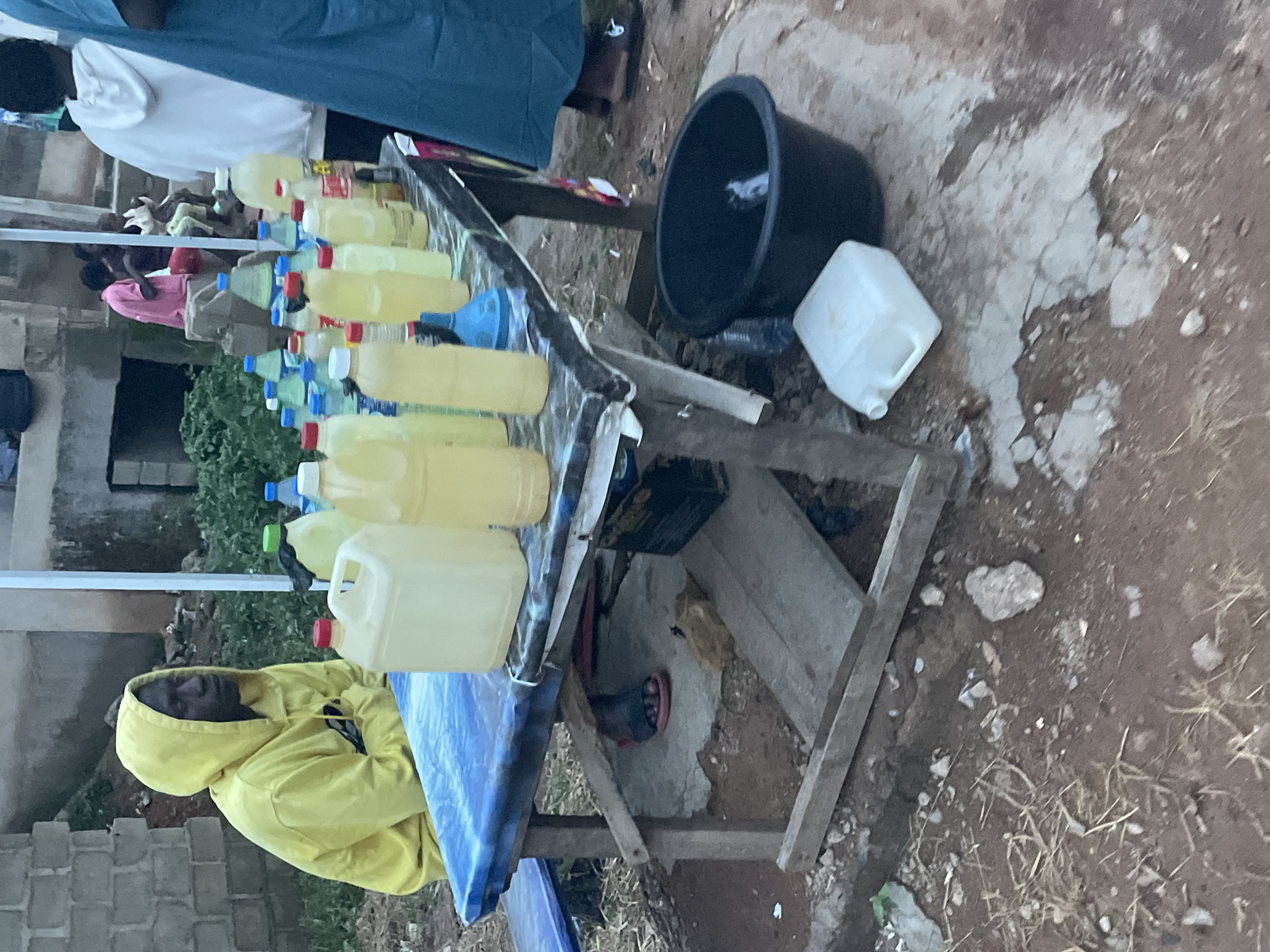 The image depicts an outdoor scene in Durumi, Nigeria. A makeshift wooden table displays numerous plastic bottles and containers filled with a yellow liquid, possibly cooking oil or fuel, suggesting a small-scale vendor operation. A man wearing a yellow hooded sweatshirt is seated on a chair near the table, seemingly resting or waiting for customers. A black plastic tub and a white plastic container are placed at the end of the table. The ground is dirt and partially paved with concrete. Other people are in the background.