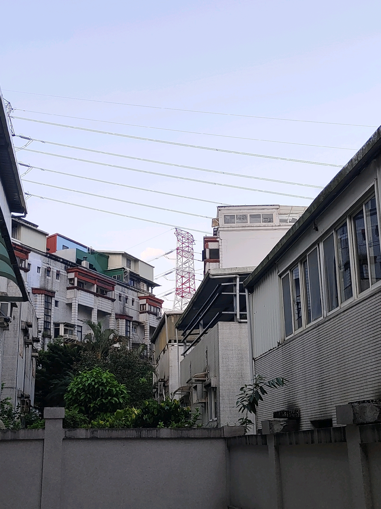 The image depicts a street scene in New Taipei City, Taiwan, focusing on residential buildings and overhead power lines. In the foreground, a grey concrete wall stretches across the frame, with a smaller wall or planter box on top filled with dense green foliage. Behind this, several multi-story residential buildings are visible, characterized by varied architectural styles, colors, and window arrangements. Some buildings have balconies and visible air conditioning units.
Dominating the background and slightly to the right is a large, red and white lattice-style electricity pylon. Numerous parallel power lines crisscross the sky above the buildings, extending from left to right across the upper portion of the image. The sky is overcast with a pale blue hue, suggesting a cloudy day. The scene appears to be an urban residential area with significant electrical infrastructure. No people are visible in the frame.