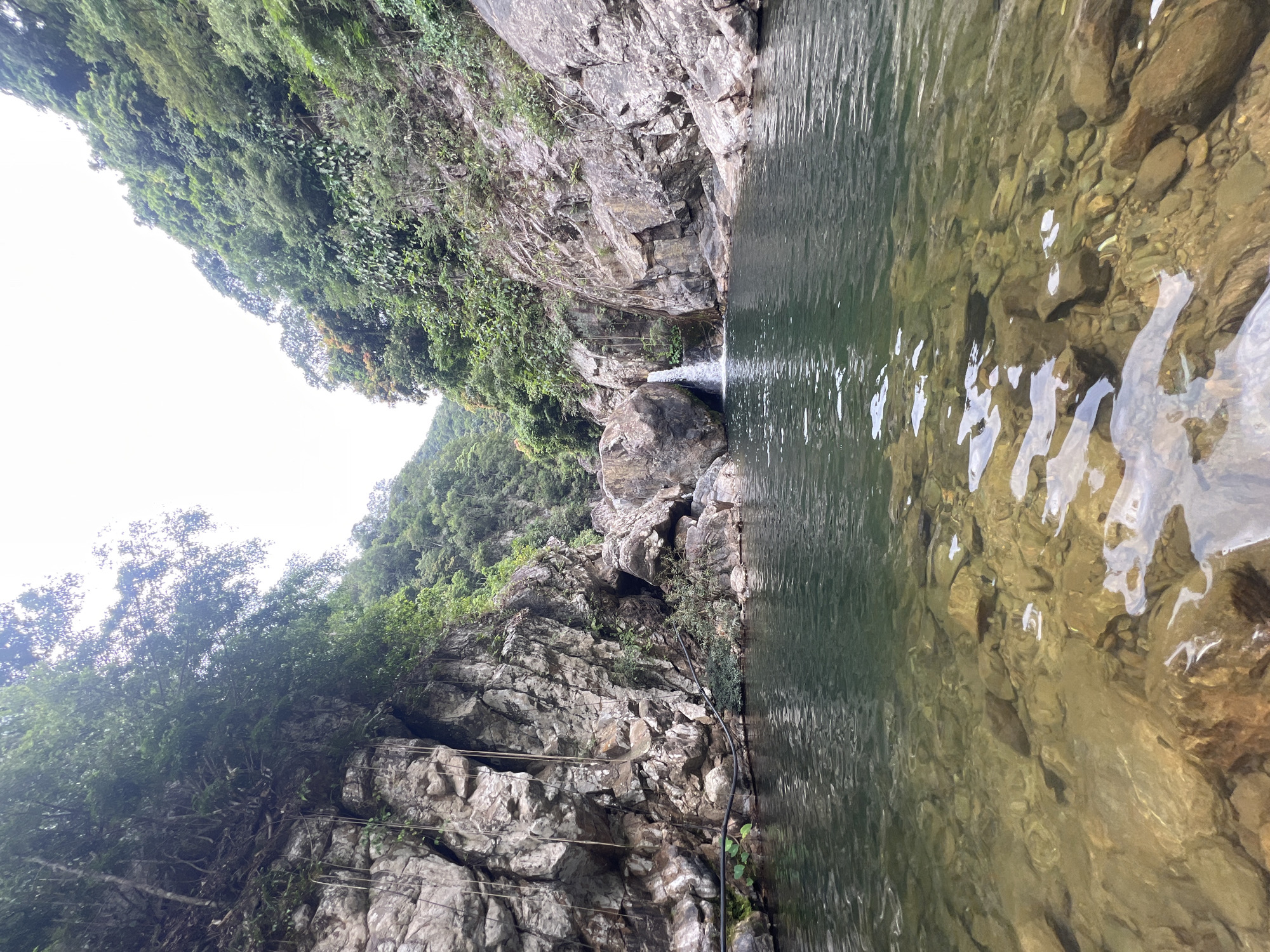 The image shows a natural scene of a waterfall cascading into a clear pool of water, surrounded by lush green vegetation and rocky cliffs. The foreground features the transparent water revealing the rocky bottom, with sunlight reflecting off the surface. The middle ground showcases the pool of water and the waterfall, which is relatively small. The background consists of steep, verdant hillsides and rocky formations covered in dense foliage. The overall environment suggests a secluded and natural setting, likely a destination for outdoor recreation. This scene is located at the Suoi Nghe Waterfall in Da Nang, Vietnam. There are no people or animals visible in the image.