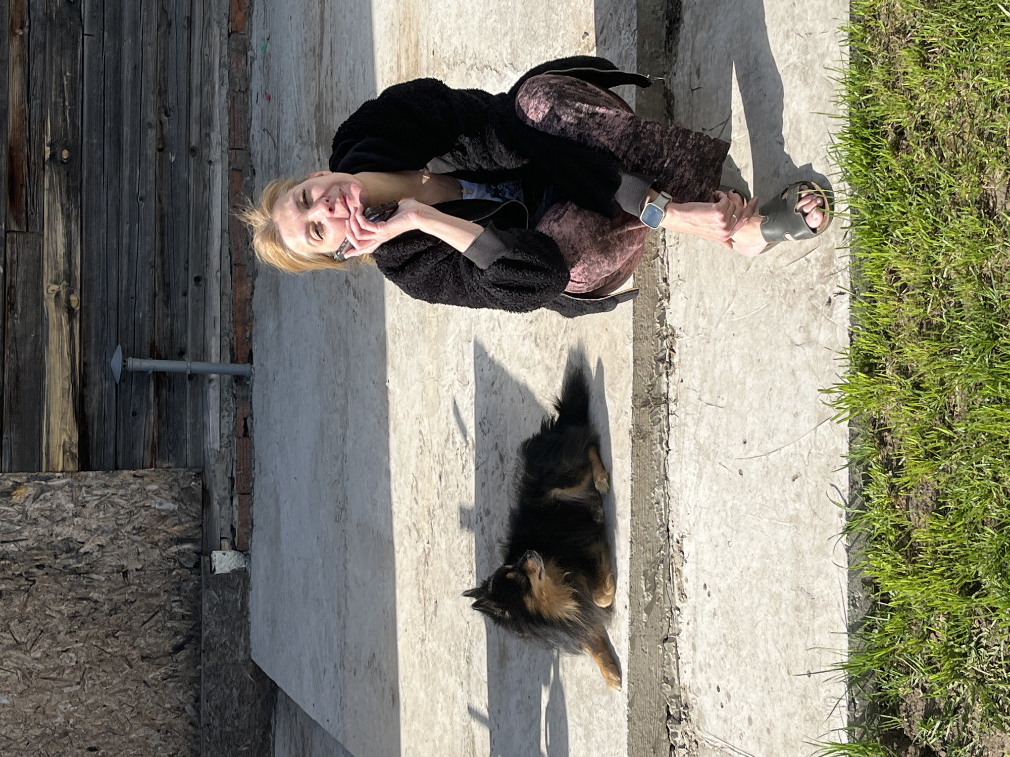A woman is sitting on a concrete step outdoors, holding a phone to her face. She has blonde hair and is wearing a black jacket and patterned pants, and sandals. A dog lies in the sun next to her. To the left of the concrete is an aged wooden wall. To the right of the concrete is green grass. There is no evidence to support that this image was taken in Izhevsk, Russia.