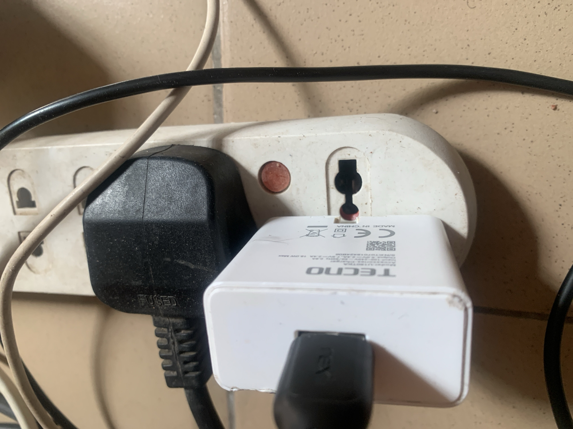 A white, multi-socket extension cord is mounted on a wall. A black, three-pin plug is inserted into one of the sockets. A white TECNO phone charger is plugged into another socket of the extension cord. The charger has a black USB cable attached to it. The scene appears to be an indoor electrical outlet, possibly in a residential or commercial setting in Jauro Yinu, Nigeria. The extension cord and surrounding wall show signs of dust and wear.