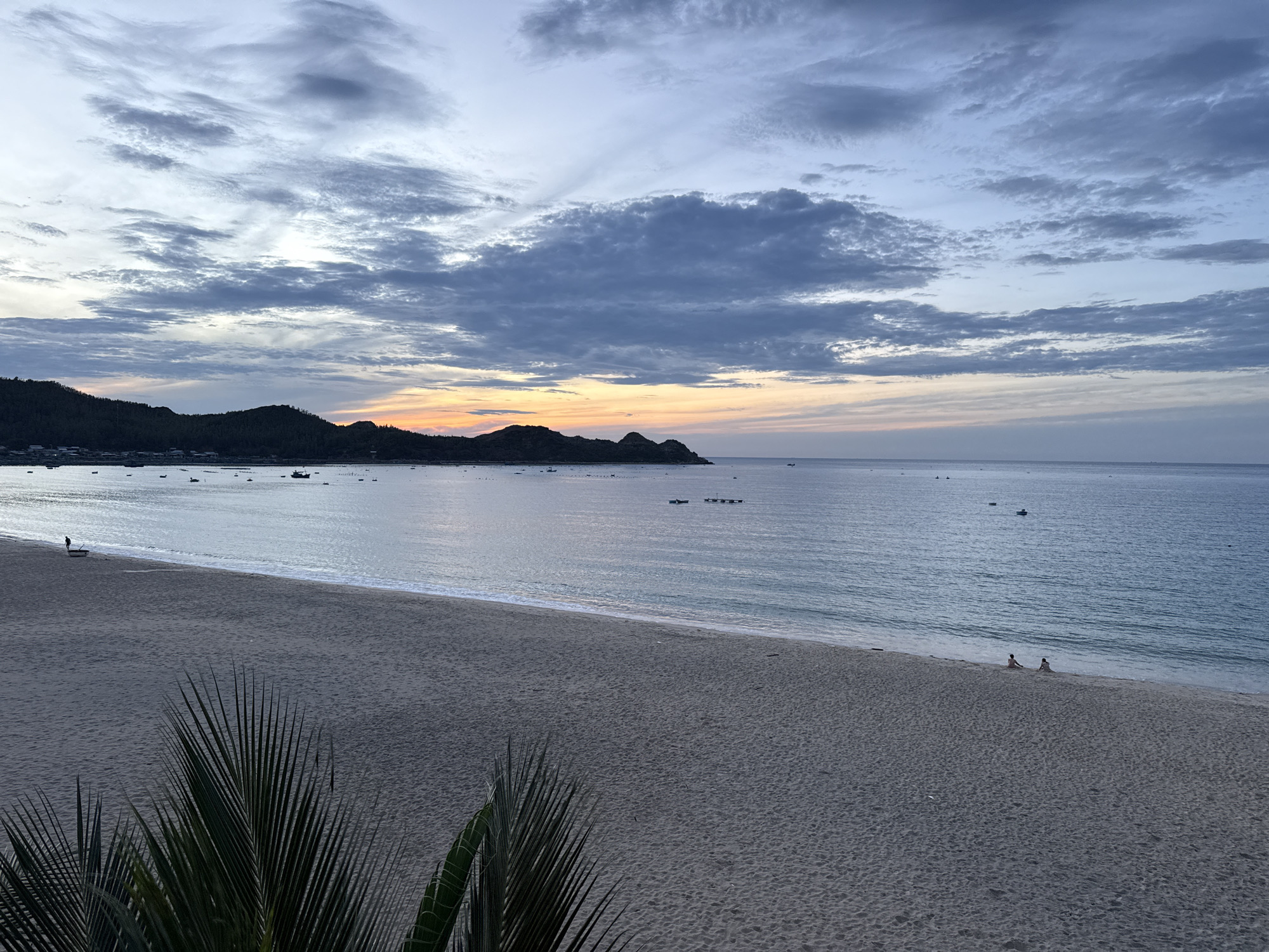 An aerial, high-angle, wide shot captures a serene beach scene in Song Cau, Vietnam, as dusk approaches. The sky is a canvas of muted blues and grays, with textured clouds streaked across it, hinting at the fading light of sunset. The horizon line is clearly defined by the meeting of the sea and sky. In the distance, a range of dark, mountainous islands with scattered lights from a settlement dot the coastline. The sea is a calm, vast expanse of blue, reflecting the sky and dotted with numerous small boats. The foreground is dominated by a wide, sandy beach, appearing dark and textured due to the low light. On the wet sand near the water's edge, two small figures are seated, facing the sea. In the very foreground, the fronds of palm trees, partially obscuring the view, frame the bottom of the image.