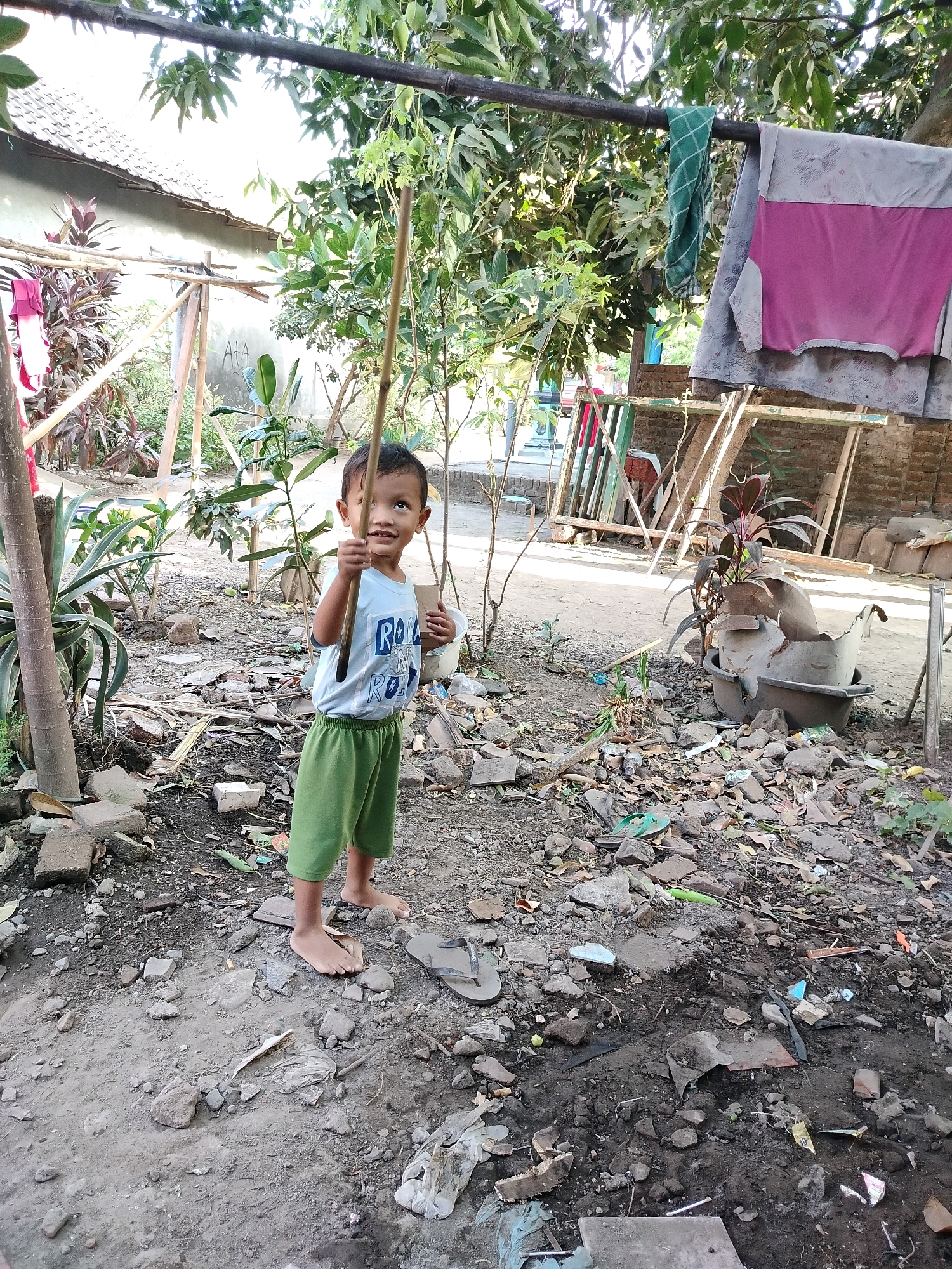 In a yard in Beloh, Indonesia, a young boy stands holding a bamboo stick. He is wearing a light blue t-shirt with white lettering and green shorts. The ground is covered in dirt, rocks, and debris, including broken pottery and discarded plastic. To the left of the boy, a house with a tiled roof is visible. In the background, laundry hangs from a clothesline, and trees provide shade. The overall scene depicts a simple outdoor environment.