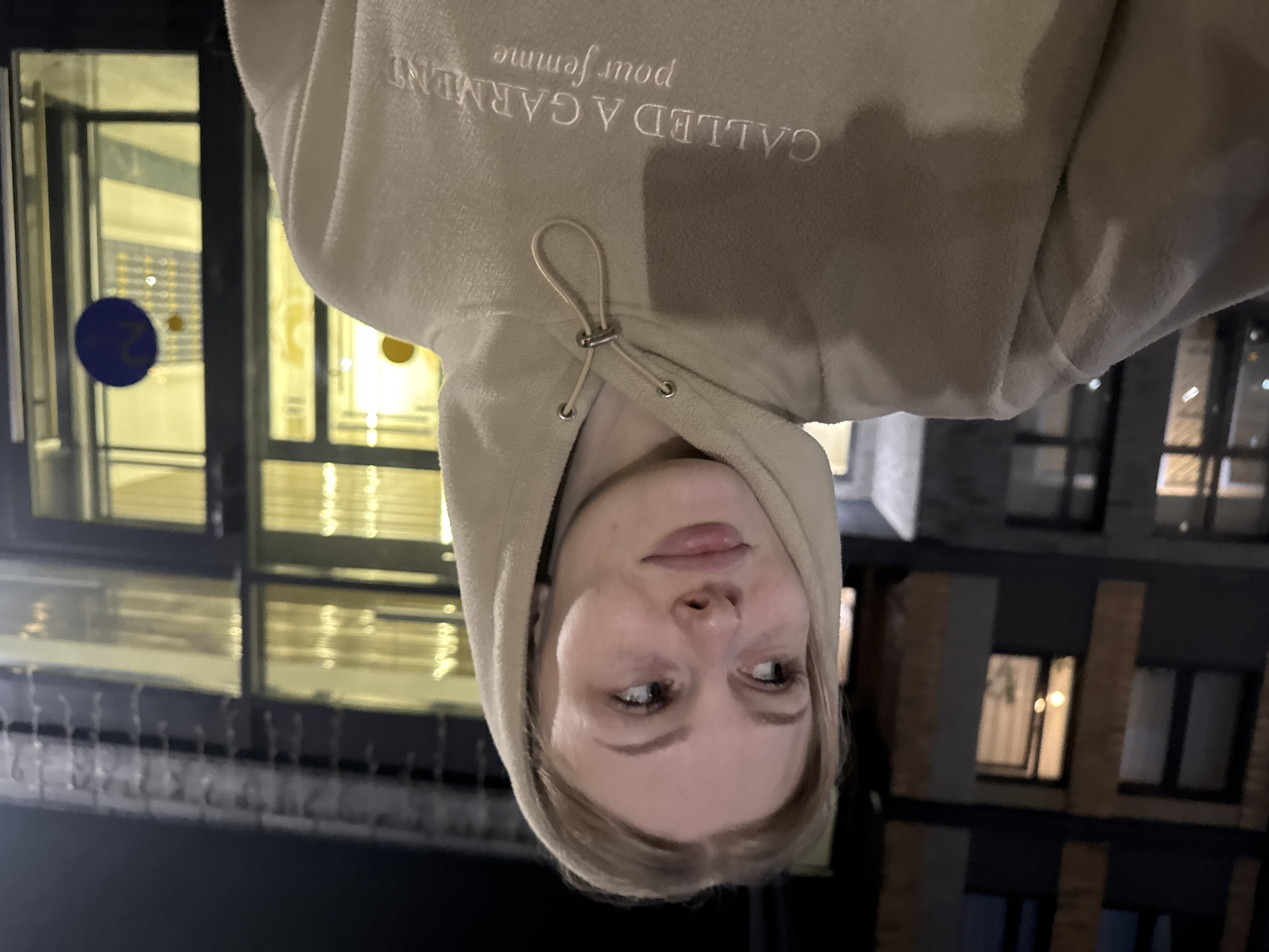 A young woman with blonde hair is standing outside at night, in what appears to be an urban environment. She is wearing a light brown hoodie. The background shows the facade of a building with illuminated windows and some sort of entrance. A blue circular sticker with the number '2' is visible on the glass door. The lighting suggests it is nighttime. The text on the hoodie reads "CALLED A GARMENT pour femme". Based on visual cues and the presence of similar architecture and building design, this scene is likely located in the city of Perm, Russia.