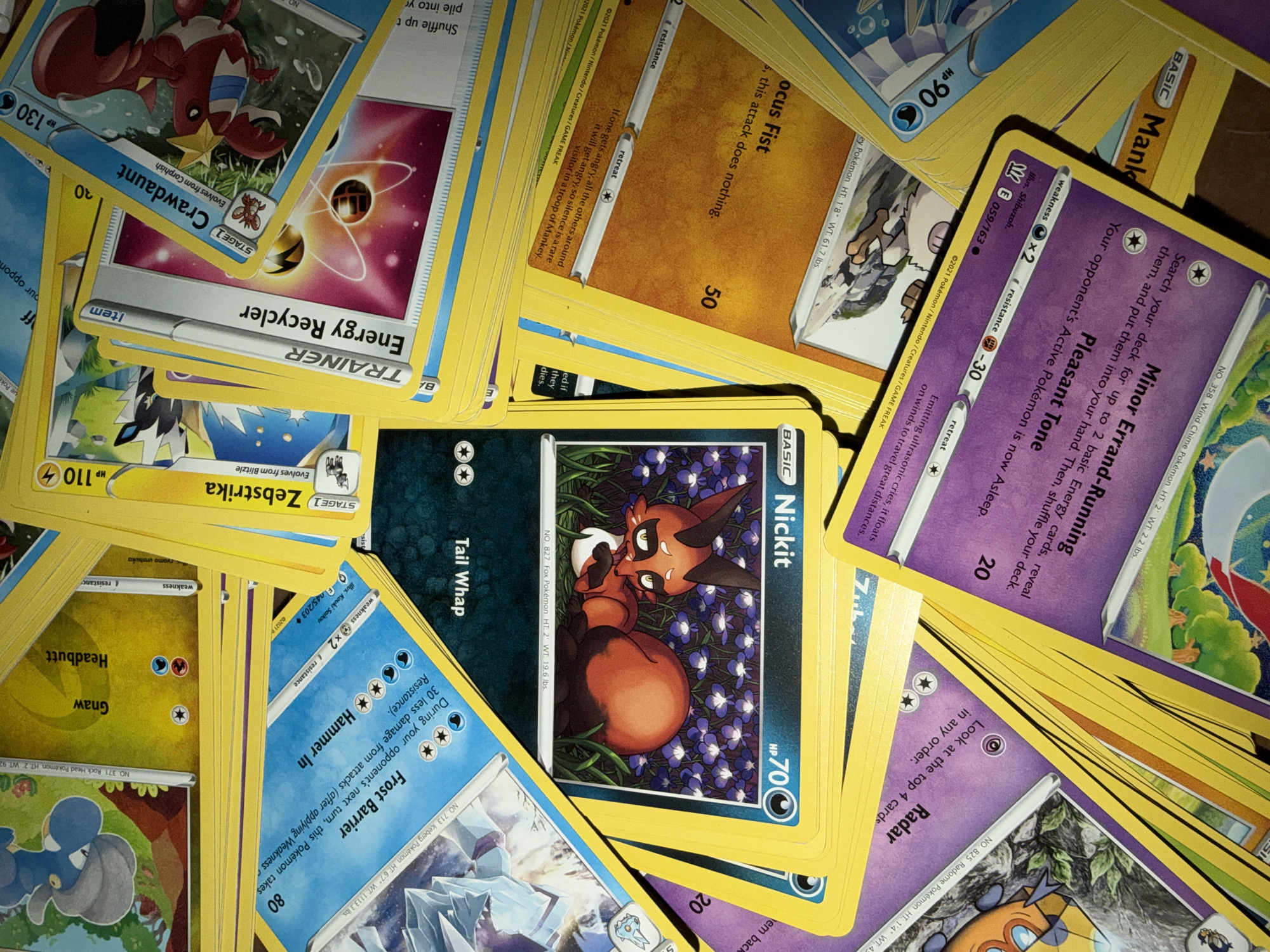 The image shows a pile of Pokémon trading cards scattered on a surface, possibly a table or floor. The cards are colorful and depict various Pokémon characters, attack names, and statistics. Some of the visible cards include "Frost Barrier," "Hammer In," "Tail Whap," "Nickit," "Energy Recycler," "Crawdaunt," "Zebstrika," "Radar," "Minor Errand-Running," and "Pleasant Tone." The lighting suggests it is indoors, with shadows cast across the cards. The specific location within Portland, United States of America, cannot be determined from the image alone as there are no identifiable landmarks or environmental cues.
