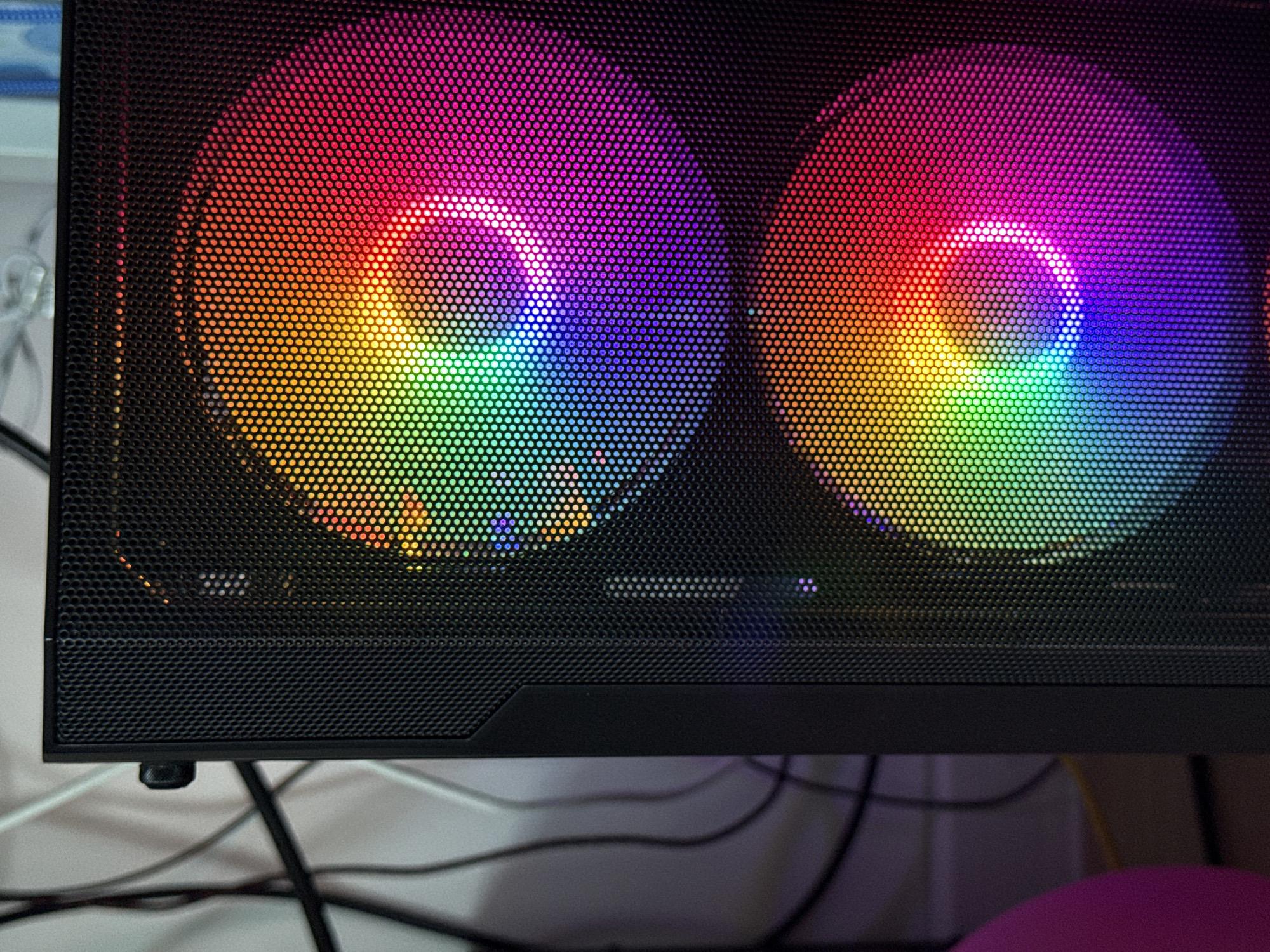 This image presents a close-up, static view of the front or side panel of a black computer tower, situated in what is understood to be the city of Targovishte, Bulgaria. The panel features a fine, black, perforated mesh grille. Through this grille, two circular cooling fans are prominently visible, each emitting vibrant, multi-colored light. The illumination transitions through a spectrum from green and yellow to orange, red, magenta, and blue, creating a distinct rainbow effect within the circular fan areas. Brighter, concentrated points of light are discernible towards the center of each fan, where a partial illuminated ring is formed around the fan hubs. To the immediate left of the mesh panel, a solid black structural element of the computer case is present, from which a small, round black knob or button protrudes near the top. The background, largely out of focus, reveals a light-colored wall against which multiple tangled black and white cables are arranged. A blurry, indistinct pink object is partially visible in the lower-left corner of the frame. No individuals or specific actions are depicted.