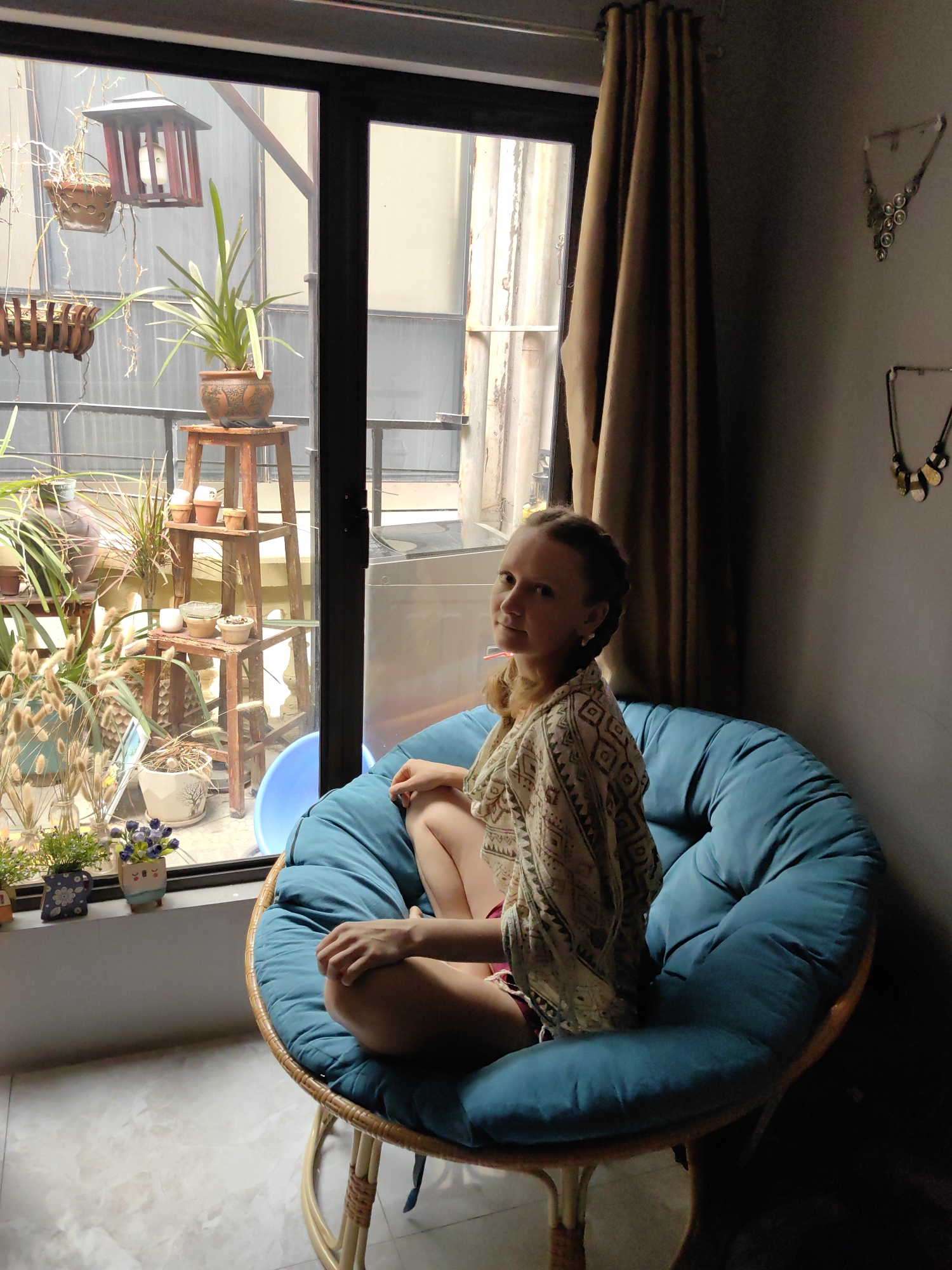 A young woman with braided hair and fair skin sits cross-legged in a large, round papasan chair with a teal-blue cushion and rattan frame. She is wearing a light-colored, patterned shawl or top and looks directly at the viewer with a calm expression.
The chair is positioned indoors, adjacent to a large window framed in black, which provides the primary light source for the room. Through the window, an outdoor balcony or patio space is visible, densely decorated with various potted plants on multi-tiered wooden stands and in hanging baskets. A wooden lantern-style birdhouse hangs from above, and a light blue plastic basin sits on the ground. A washing machine is partially visible on the balcony, and beyond it, the corrugated facade of another building can be seen, typical of an urban setting like Hà Nội.
Inside, the windowsill features small potted plants and decorative items. To the right of the window, a brown curtain is drawn, and on the adjacent grey wall, two distinct necklaces are displayed as decor. The floor is tiled. The scene is illuminated by natural, indirect daylight, suggesting it is daytime. There is no visible text in the image.