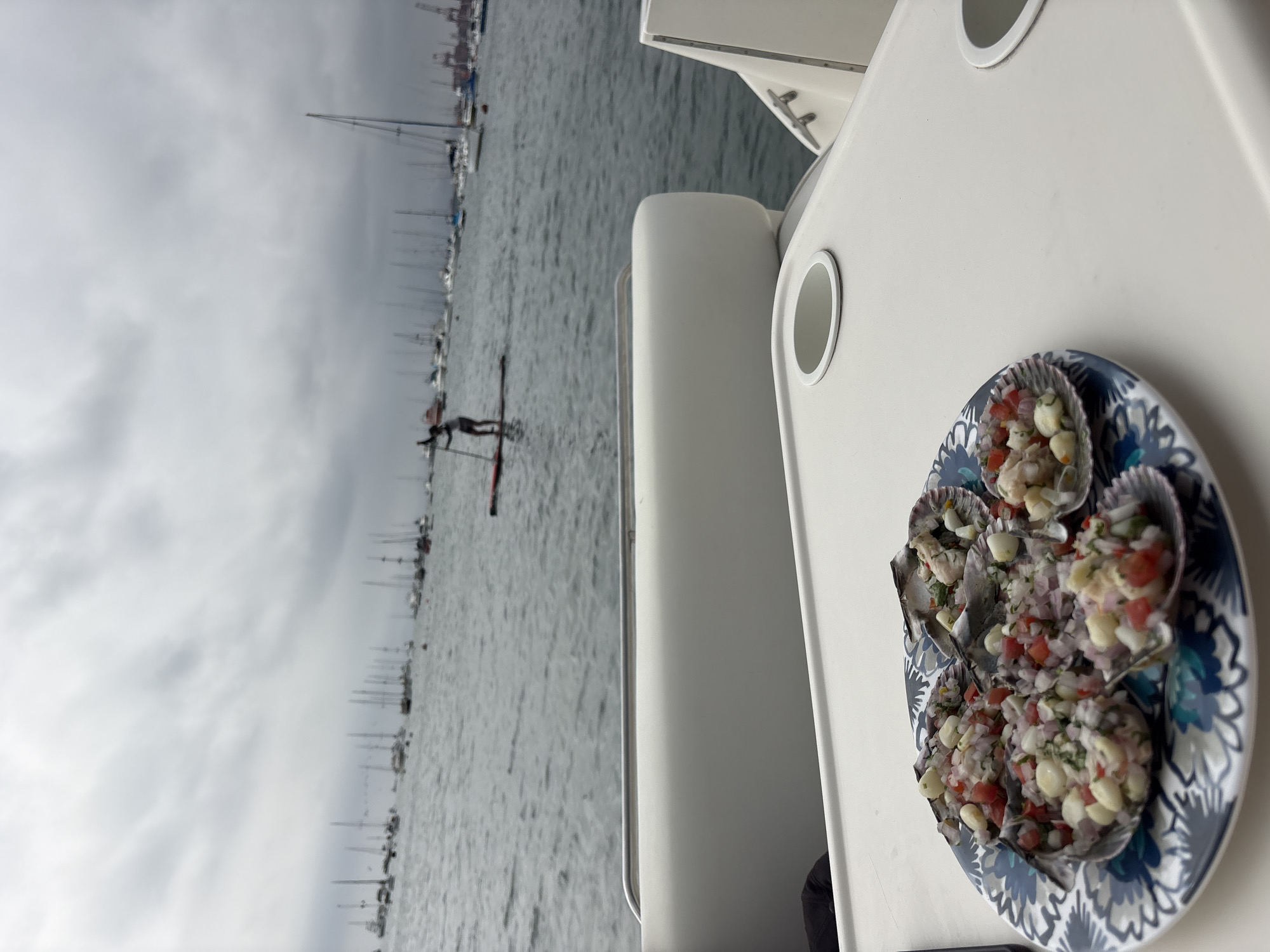 In La Punta, Peru, a plate of scallops prepared as ceviche sits on a white table on a boat. The ceviche is topped with corn and diced tomatoes and onions. In the background, a person is paddleboarding on the water, with numerous sailboats anchored in the harbor. The sky is overcast.