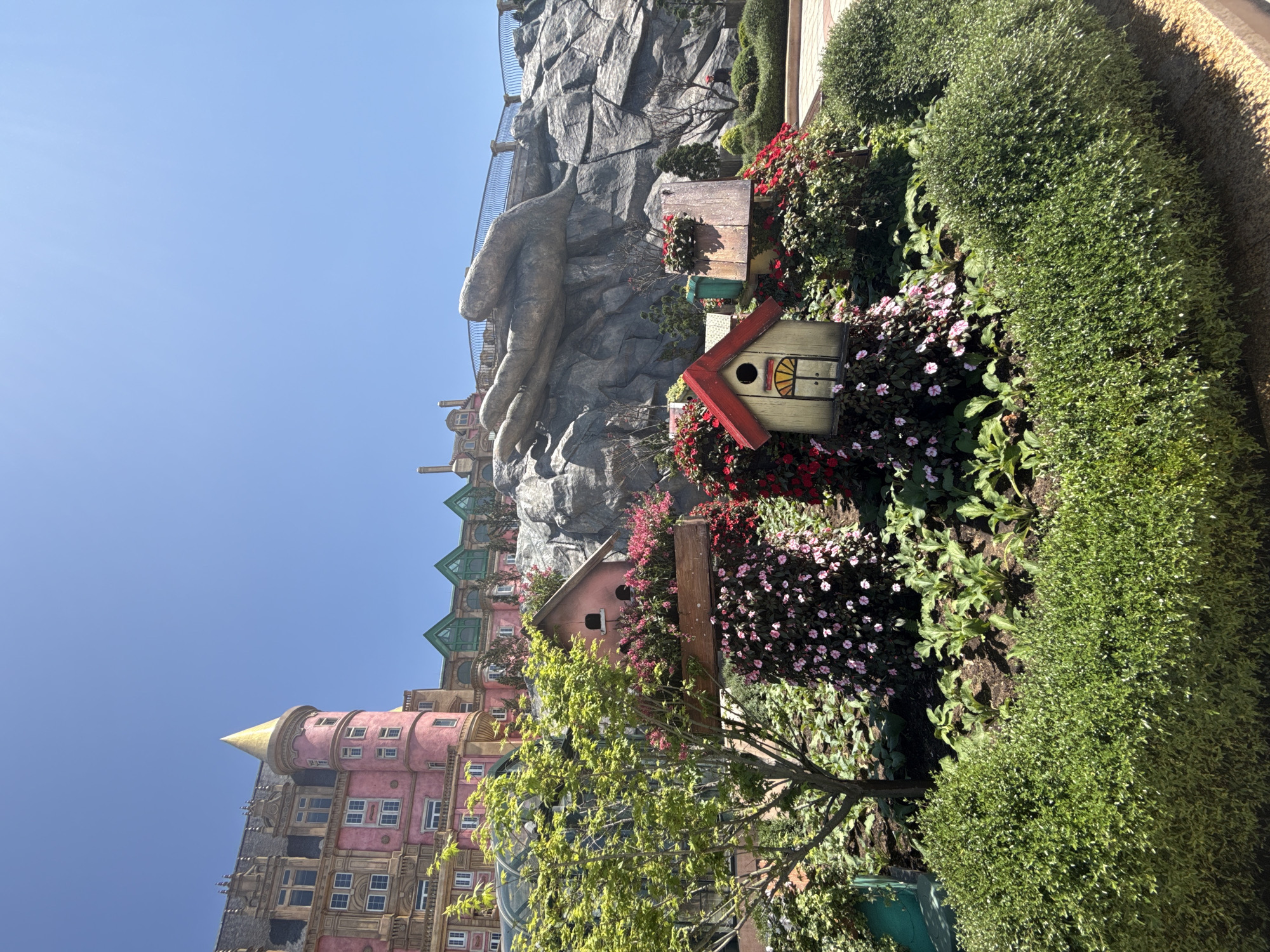 The scene is a vibrant landscape in Đà Nẵng, Vietnam, featuring a fantastical architectural ensemble. A multi-story building, colored in hues of pink, yellow, and green, is partially visible, with a golden conical turret projecting towards a clear blue sky. A massive sculpted stone hand emerges from a rocky outcrop, appearing to support a pedestrian bridge above. In the foreground, there's a landscaped garden with lush greenery, flower beds filled with red and pink blooms, and two birdhouses nestled among the plants. The garden is bordered by a neatly trimmed hedge.