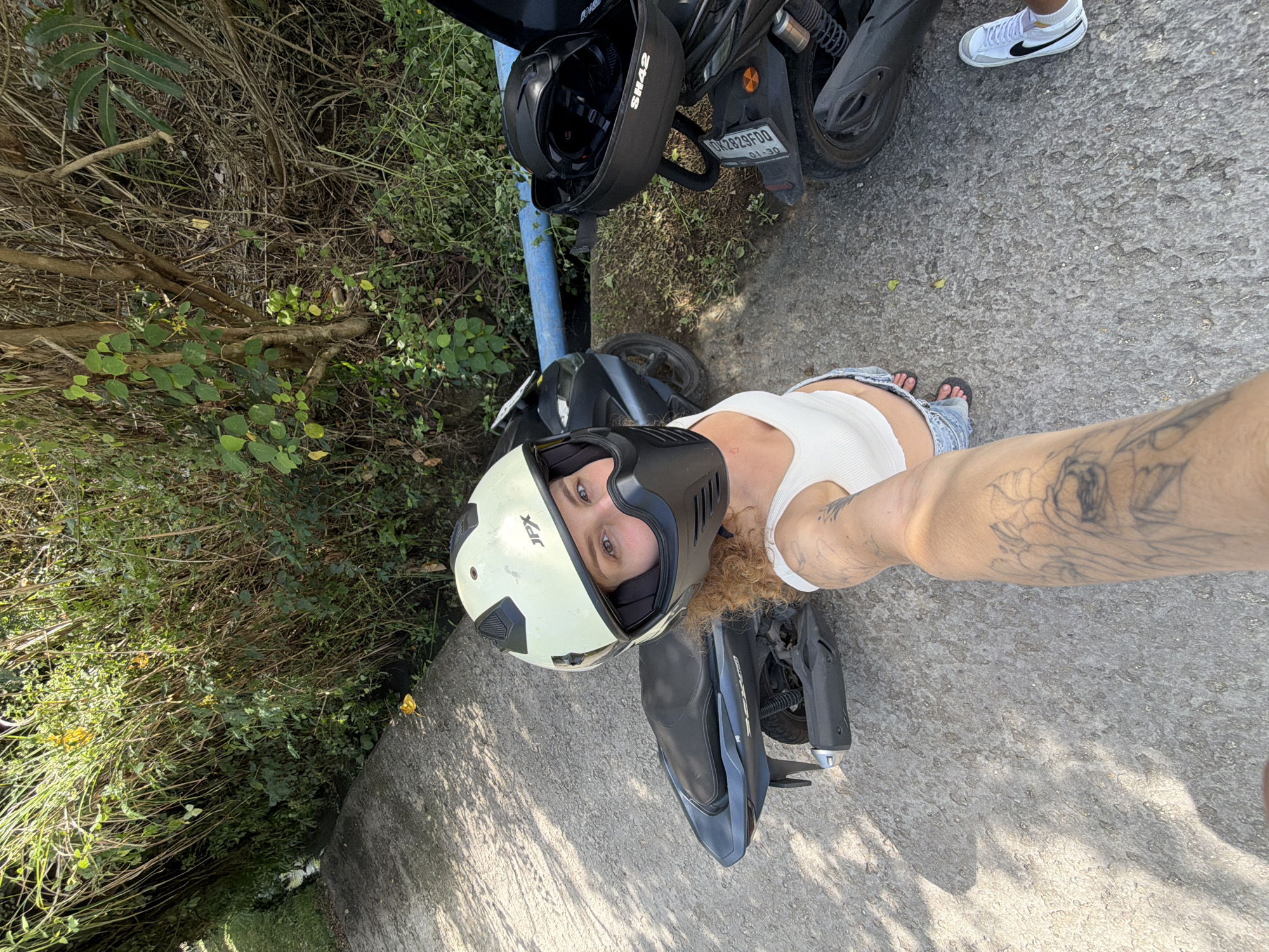 In Kediri, Indonesia, a woman with curly blonde hair, wearing a white tank top and denim shorts, poses for a selfie while riding a scooter. She has a light-colored helmet with a black chin guard. The scooter is dark gray, and another scooter with a black helmet is parked nearby. The scene is on a paved road bordered by lush green vegetation and trees. Sunlight casts shadows on the road.