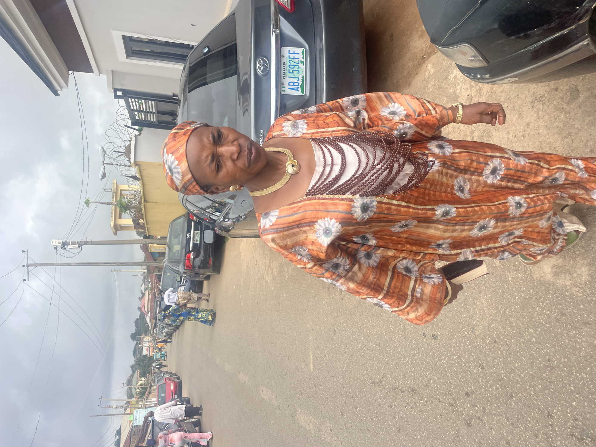 The image depicts a street scene in Jos, Nigeria. The foreground is dominated by a woman wearing an orange and white patterned dress and headscarf, along with several beaded necklaces and gold jewelry. She is holding a book in her left hand. To her right, a dark sedan is parked, displaying a Nigerian license plate "ABJ 592FF". The street is paved and lined with parked cars and buildings on both sides. Several other individuals in traditional attire are visible in the background, walking along the street and near parked vehicles. The sky is overcast.