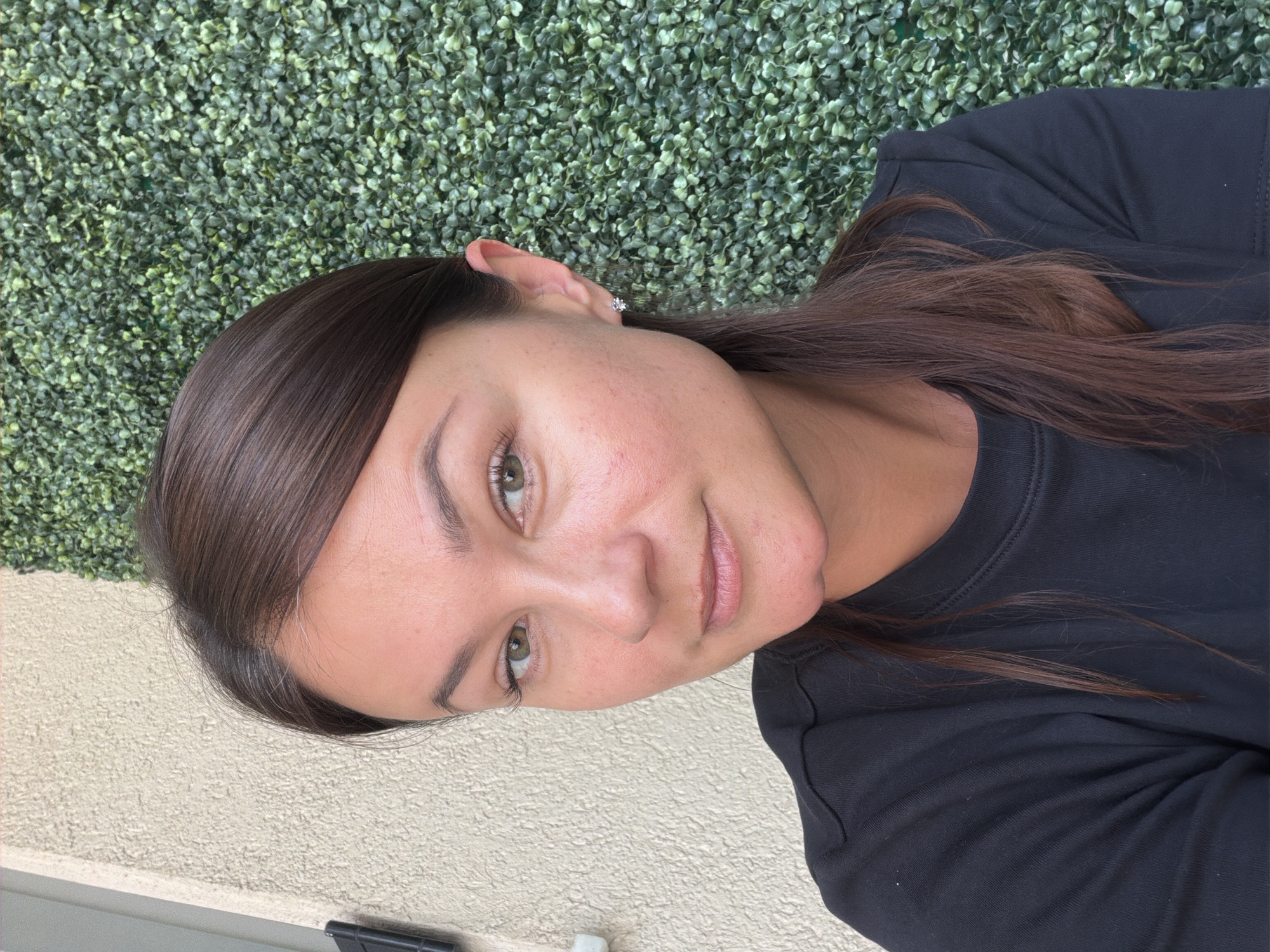 The media file is a portrait of a young woman with long, dark brown hair. She is wearing a black crew neck shirt. Her hair is pulled back on one side, revealing her ear which has a small stud earring. Her eyes are a light green color, and she has light-colored eyebrows. She has light skin with some redness on her cheeks. Her lips are slightly parted. She is positioned in front of a textured green wall that resembles hedges or artificial foliage. To the left of the green wall, there is a cream-colored textured surface, possibly a wall or building exterior. The lighting suggests it is daytime, and the overall scene appears to be outdoors in a casual setting. There is no visible text in the media file. The orientation of the image is landscape, with the woman's head on the left side of the frame.