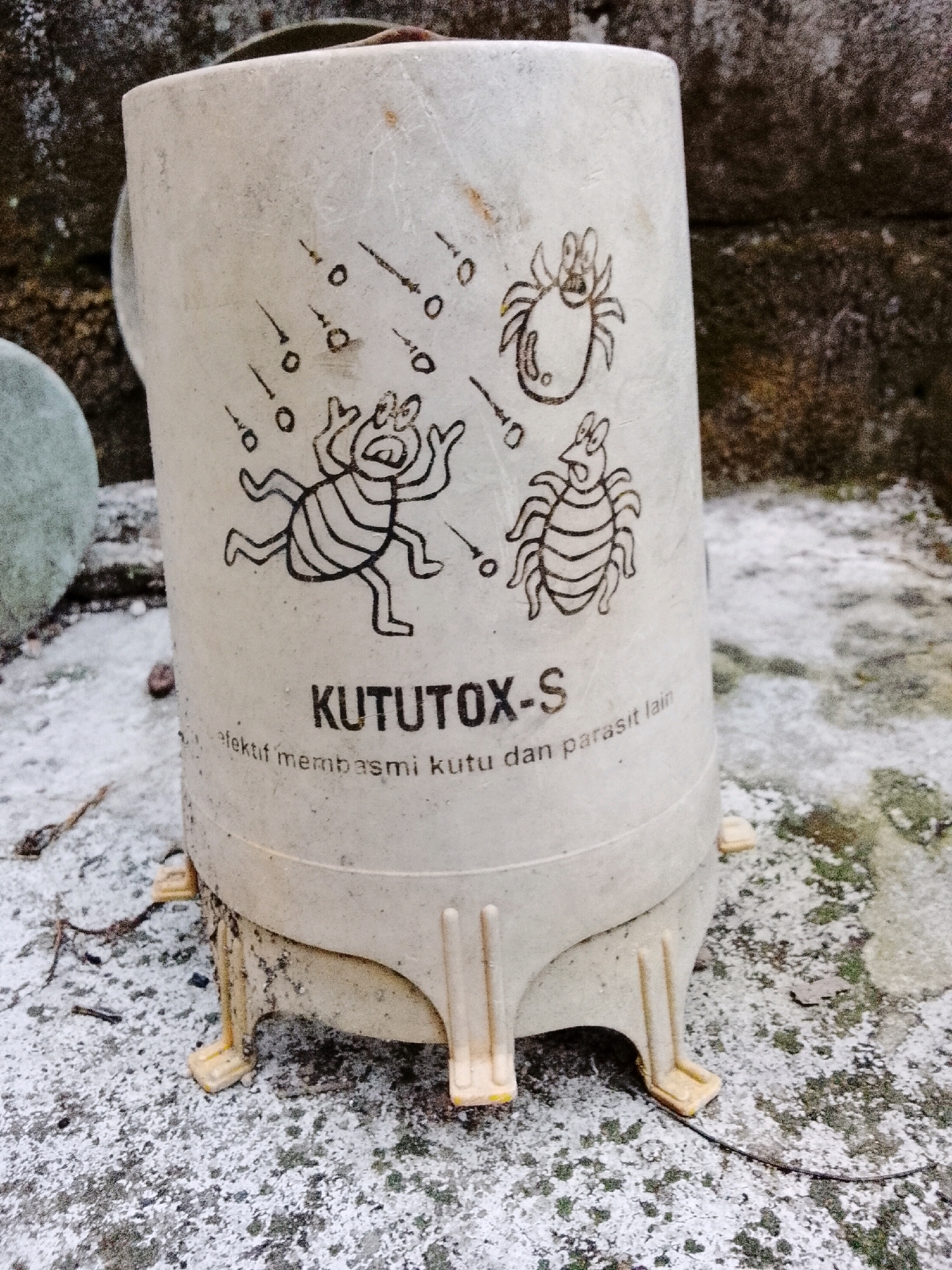 A weathered, off-white cylindrical container with cartoonish drawings of insects and accompanying text sits on a textured, greyish-white surface that appears to be speckled with green moss and dirt. The container, which has three visible yellow legs supporting it, is positioned slightly off-center to the right of the frame.
The drawings on the container depict three stylized insects: one in the upper right corner with multiple legs and a wide, open mouth, seemingly reacting to falling black dots and streaks depicted above it. To its left, another insect with a similar body shape and six legs is shown. Below these, a larger, multi-striped insect with arms raised in apparent distress is illustrated, also with falling dots and streaks surrounding it.
Beneath the drawings, the text "KUTUTOX-S" is prominently displayed in black capital letters. Below this, in smaller print, is the phrase "efektif membasmi kutu dan parasit lain".
The surface the container rests on is uneven and appears to be outdoors, possibly a stone or concrete surface. Small debris, including twigs and leaf fragments, is scattered on the surface. In the background, a rough, dark, textured wall is visible. A portion of a round, light-colored object, possibly another container or a plant, is visible on the far left edge of the frame, partially obscured. The overall lighting suggests daylight. The scene is devoid of any people or human interaction.
The location is identified as Padang Dalom, Indonesia.