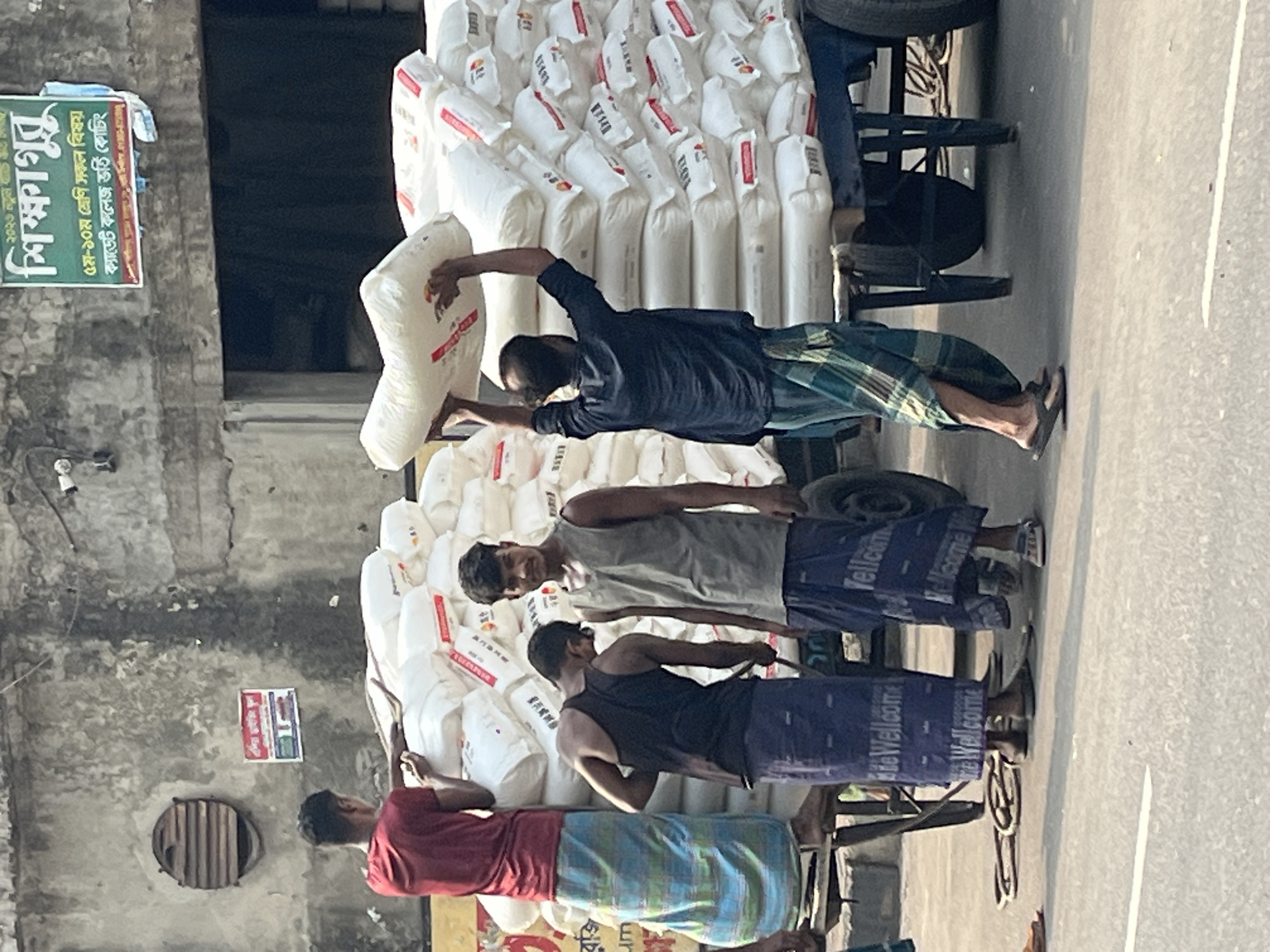 The image depicts a bustling scene on what appears to be a street in Dhaka, Bangladesh. Four men are loading large white sacks, likely containing goods such as rice or flour, onto what seems to be modified rickshaws or carts. The men are wearing a mix of casual clothing, including shirts and lungis (traditional wraparound garments).
The sacks are stacked high on the carts, suggesting a substantial load. The environment includes buildings lining the street, one of which features a sign with Bengali script.