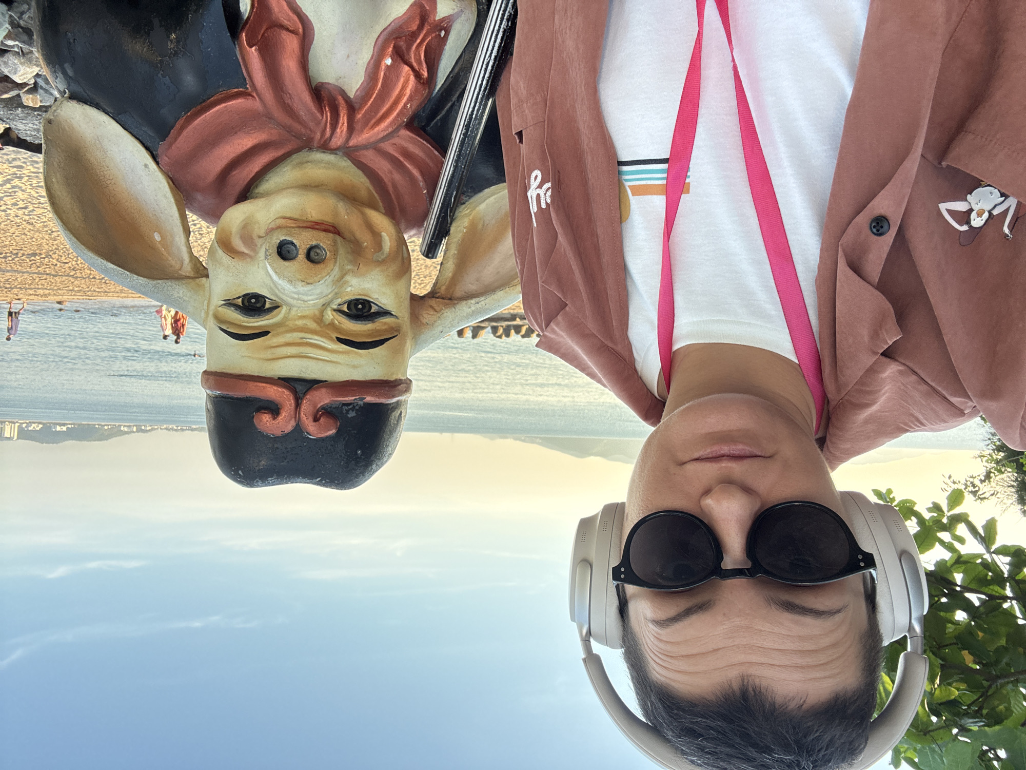 A woman with short dark hair, wearing headphones, sunglasses, and a pink lanyard around her neck, stands on a beach in Nha Trang Ward, Vietnam, next to a statue of a pig. The woman is wearing a light pink open shirt over a white t-shirt with a colorful stripe design. The pig statue is wearing a black hat with red trim and a red scarf around its neck. The statue is made of painted material and has human-like eyes and ears. In the background, the ocean is visible, with a few people walking on the beach and distant hills or mountains. The sky is clear and blue with some wispy clouds. The lighting suggests it is late afternoon or early evening.