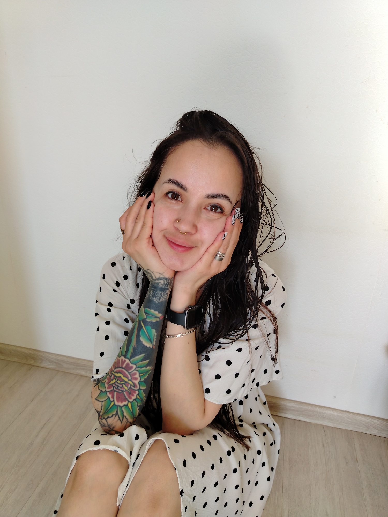 A woman with brown hair and a nose piercing is sitting on a light wood floor against a white wall. She is wearing a white dress with black polka dots. Her hands are supporting her chin, and she is smiling. Her left arm has a colorful tattoo. She is wearing a silver bracelet and a black smartwatch on her left wrist. Her fingernails have a black and white pattern.
There is no indication in the image that the location is in the city of Noviy, Russia.