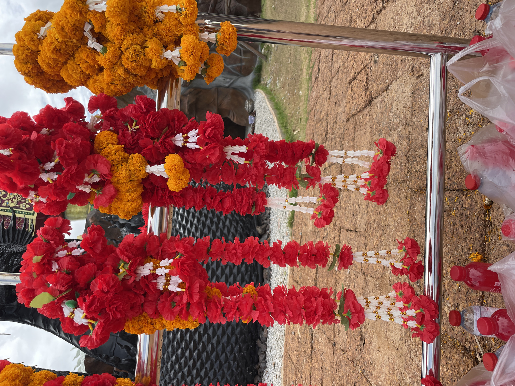 The image shows a close-up of several flower garlands hanging on a metal railing. The garlands are primarily composed of red and orange flowers, with some white and gold decorative elements interspersed. In the background, there is a glimpse of a dark, textured surface, possibly part of a statue or structure, and a dry, rocky ground area with some sparse greenery. Below the railing, several plastic bottles, some filled with red liquid, are visible, along with clear plastic bags containing more items. The overall scene suggests an outdoor location, possibly a religious or ceremonial site in the city of Bo Win Subdistrict, Thailand. The lighting indicates it is daytime.
