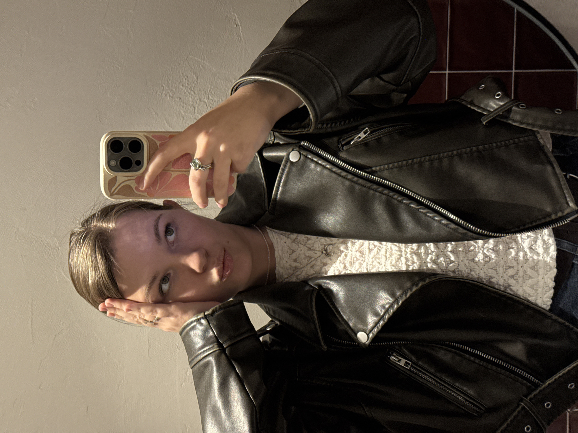 A young woman is depicted in a mirror selfie, holding an iPhone to capture her reflection. She has light brown hair, pulled back, and light-colored eyes. Her expression is neutral, and she gazes directly towards the camera. One hand holds the phone, revealing a silver-toned ring with a distinctive, possibly floral or leaf-like, design on her middle finger. The phone has a light-colored case adorned with a soft pink and brown abstract or floral pattern. Her other hand rests gently against her cheek, partially obscuring her jawline.
She is dressed in a black, biker-style leather jacket featuring numerous zippers, silver snaps, and buckled details. A white, textured top, possibly lace or a delicate knit, is visible underneath the jacket.
The background shows a plain, light-colored wall, likely beige or off-white, occupying most of the space. To the right, a section of a tiled wall with reddish-brown tiles and white grout lines is visible, suggesting an indoor setting such as a bathroom or a room with specific architectural features. The lighting is warm and directional, creating subtle highlights and shadows. There is no visible text in the image.