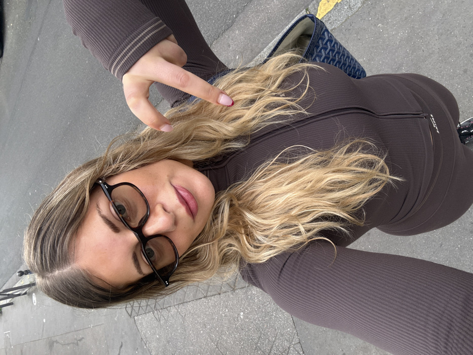 A single female subject with long, wavy blonde hair, parted centrally, is depicted in a high-angle selfie on an urban street. She wears black-framed glasses with light-tinted lenses, subtle eye makeup, and pink lipstick. Her attire consists of a ribbed, dark brown athletic-style two-piece outfit: a long-sleeved, quarter-zip top with two lighter brown stripes on the right sleeve cuff, and matching leggings. Her right hand is raised and positioned with the index finger pointing downwards; the fingernails display a French manicure, with the index finger having a distinct red tip. A dark blue tote bag with a geometric pattern is partially visible on her left side. The background features a grey concrete sidewalk lined with black metal bollards, adjacent to a darker asphalt road. A yellow curb line is visible on the far right, and a vehicle is partially discernible in the upper right background. The distinctive street furniture, particularly the bollards, indicates the location is in Paris, France.
