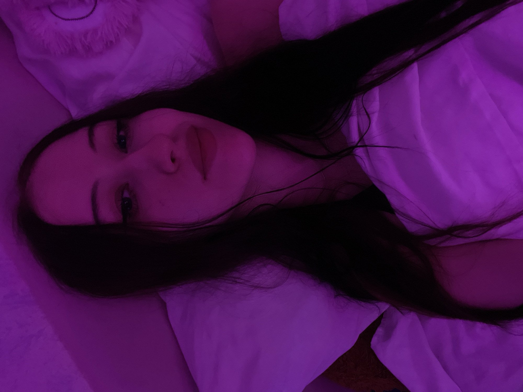 A young woman with long, dark hair lies in bed, illuminated by purple light. She is looking at the camera with her eyes open and her lips slightly parted. She is covered by a white duvet, and a white pillow is behind her head. The room appears to be dimly lit, with the purple light casting a strong glow on her face and surroundings. The overall impression is one of a relaxed and intimate moment, possibly before sleep or upon waking. The image does not provide any information about the location within the city of Votkinsk, Russia.