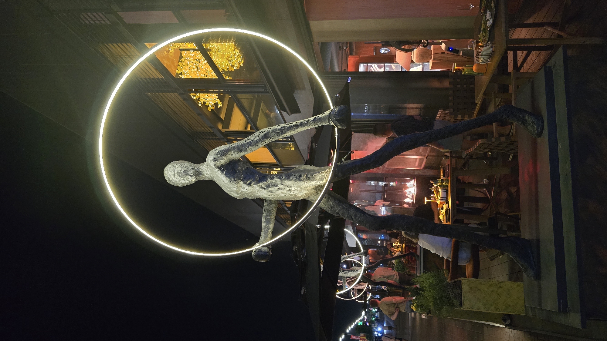 The image depicts a nighttime scene in Kyiv, Ukraine, showcasing a large, abstract humanoid sculpture at its center. The sculpture, rendered in a textured, dark metallic finish, features an elongated, slender form with disproportionately long legs and arms. Its surface appears weathered or deliberately textured to resemble organic material. A bright, circular ring of warm white light encircles the sculpture's upper torso and head, held by its outstretched left arm and passing near its right arm.
The sculpture stands on a dark, raised base, positioned on a sidewalk or pedestrian area. To the left, a series of similar sculptures with glowing rings are visible further down the street, indicating an art installation. The street is illuminated by various artificial lights, and several individuals are walking or standing in the background.
To the right of the central sculpture is the facade of a modern building with large glass windows. Through these windows, bright yellow-orange decorative lighting or an art installation resembling abstract foliage is visible on an upper floor. Below this, an outdoor dining area extends from the building, featuring several dark tables and chairs. People are seated at these tables, some engaged in conversation or dining. One table shows plates, glasses, and a bottle. Small, enclosed fire features or heaters emit a soft, orange glow near some tables. Large, dark patio umbrellas are partially open, covering parts of the dining area.
The overall environment is that of a bustling urban street at night, with a blend of artistic display, commercial establishments, and public gathering spaces. The dark sky indicates late evening or nighttime.